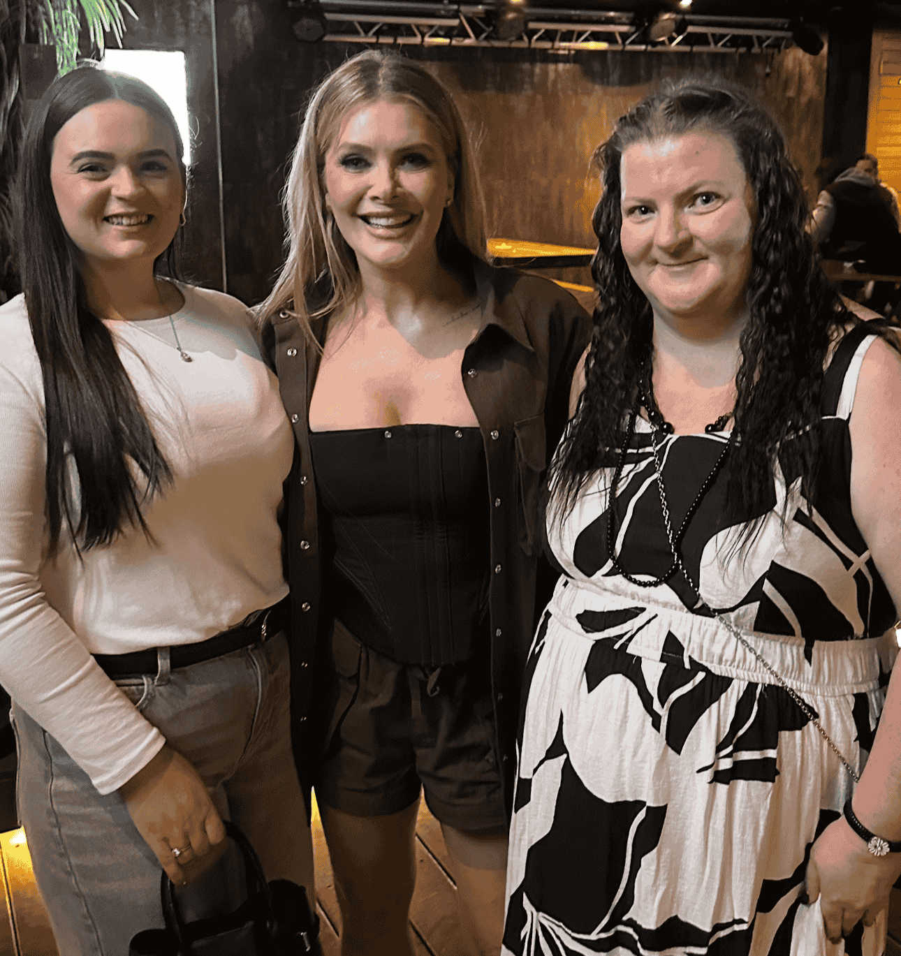 Three women smiling and standing together indoors at a social event.