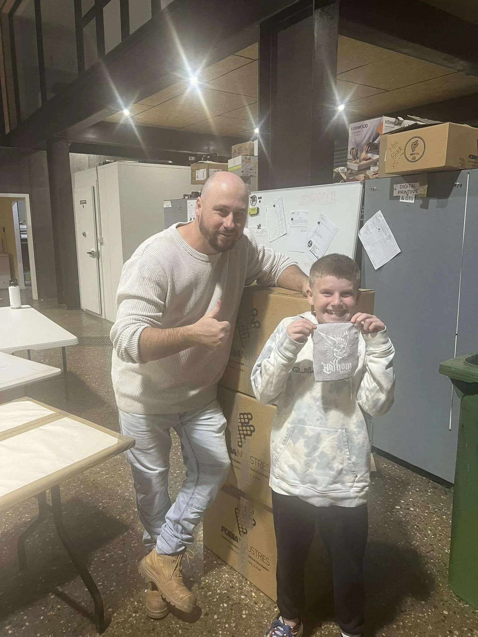 A man and a boy are smiling and posing for a photo in a room with industrial decor. The man is giving a thumbs-up and has a beard, wearing a light beige sweater, white pants, and tan boots. The boy is holding a paper or cloth with a skull design, wea