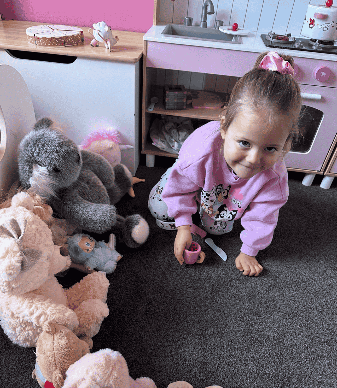 A young girl in pajamas kneeling on a carpeted floor, holding a toy cup, surrounded by stuffed animals in a play kitchen area.