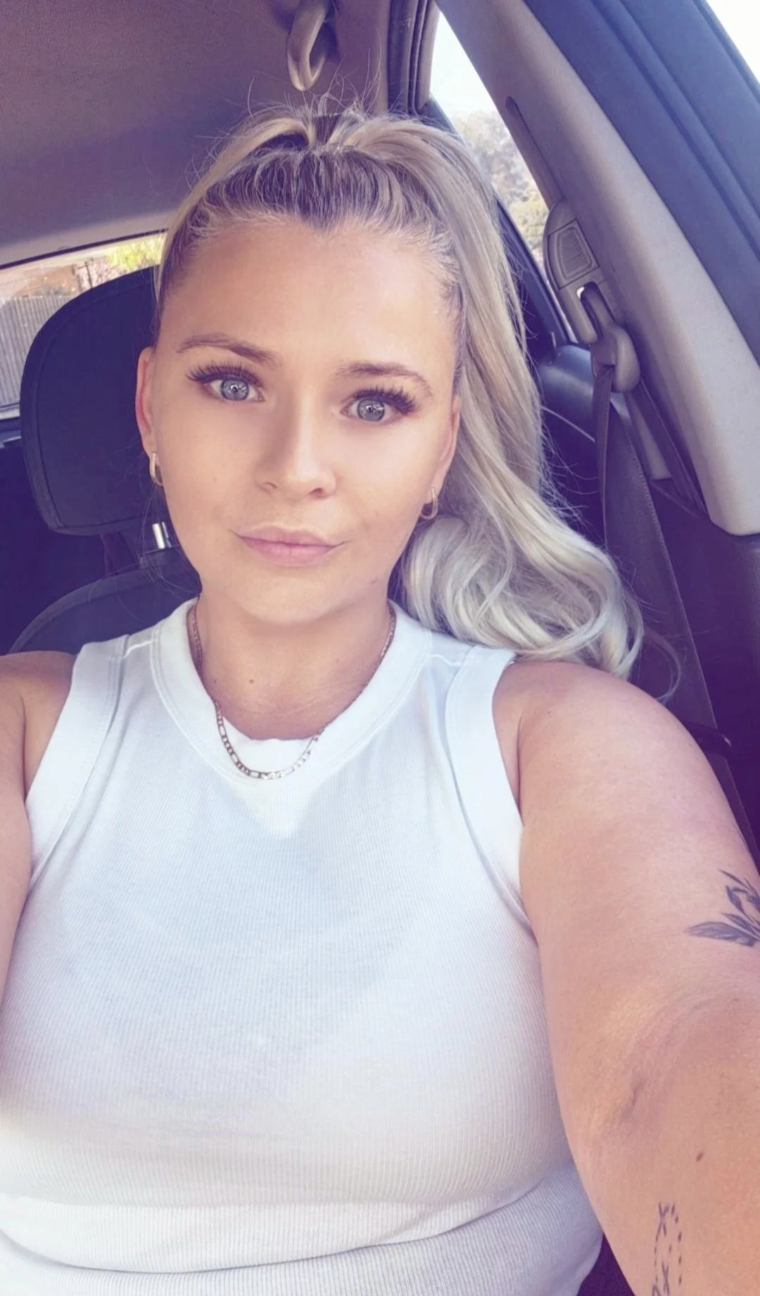 A young woman with platinum blonde hair in a high ponytail, sitting inside a car, wearing a white sleeveless top, earrings, and a chain necklace. She has blue eyes and appears to be taking a selfie.