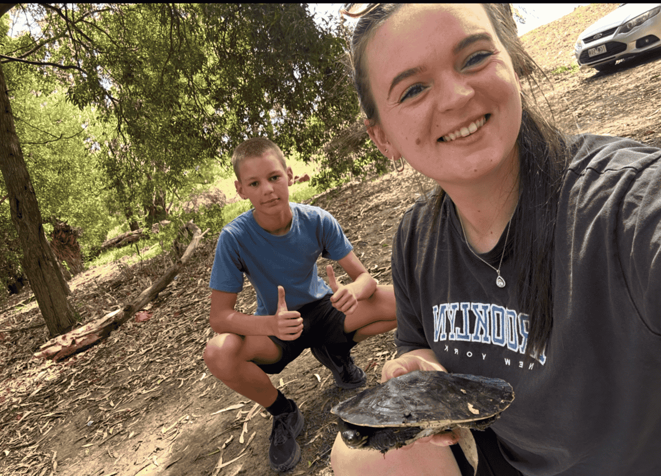 A woman and a boy outdoors in a wooded area, with the woman holding a turtle. The woman is smiling and taking a selfie, while the boy is giving a thumbs-up.