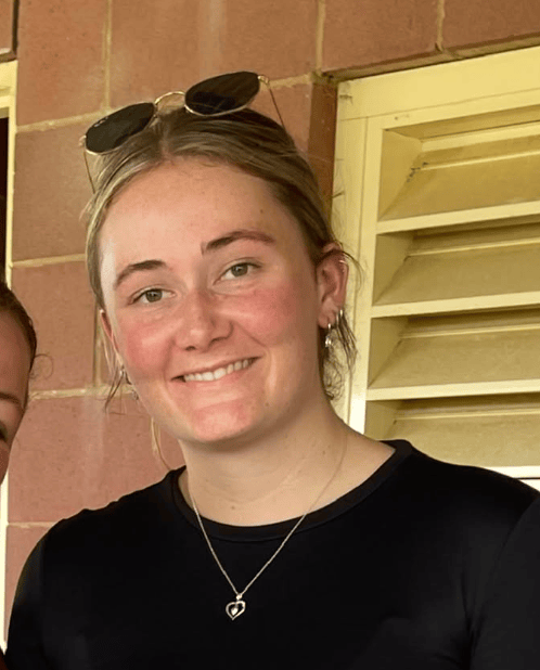 A young woman with blonde hair, sunglasses on her head, wearing a black shirt and a silver heart necklace, smiling outdoors near a brick wall and yellow window shutters.