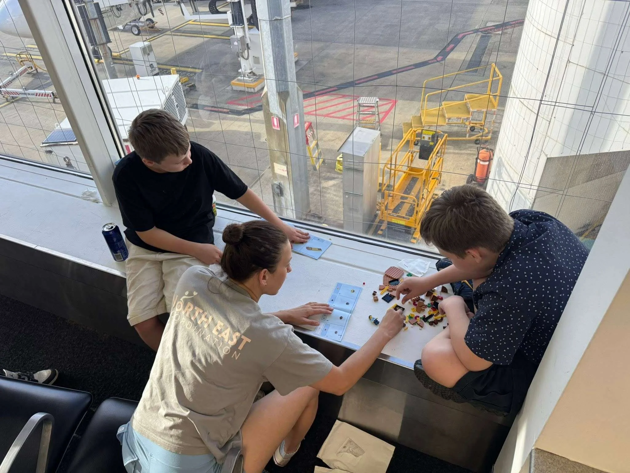 Group of four people sitting at a window ledge, playing with LEGO bricks on a table, with an airplane tarmac visible outside the window.