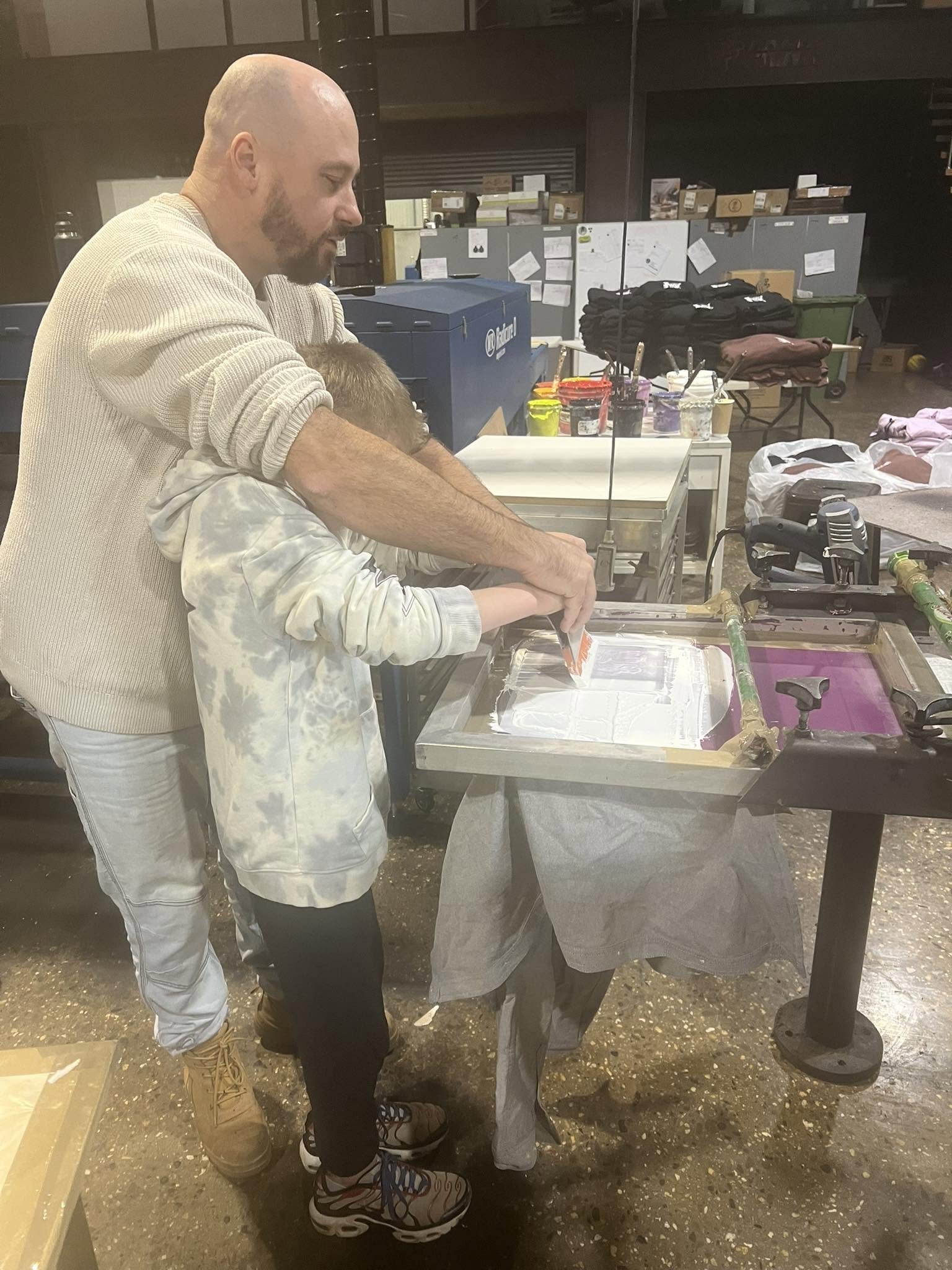 A man and a young boy working together at a table with art supplies, including paints and brushes, in an indoor setting with storage shelves and boxes in the background.