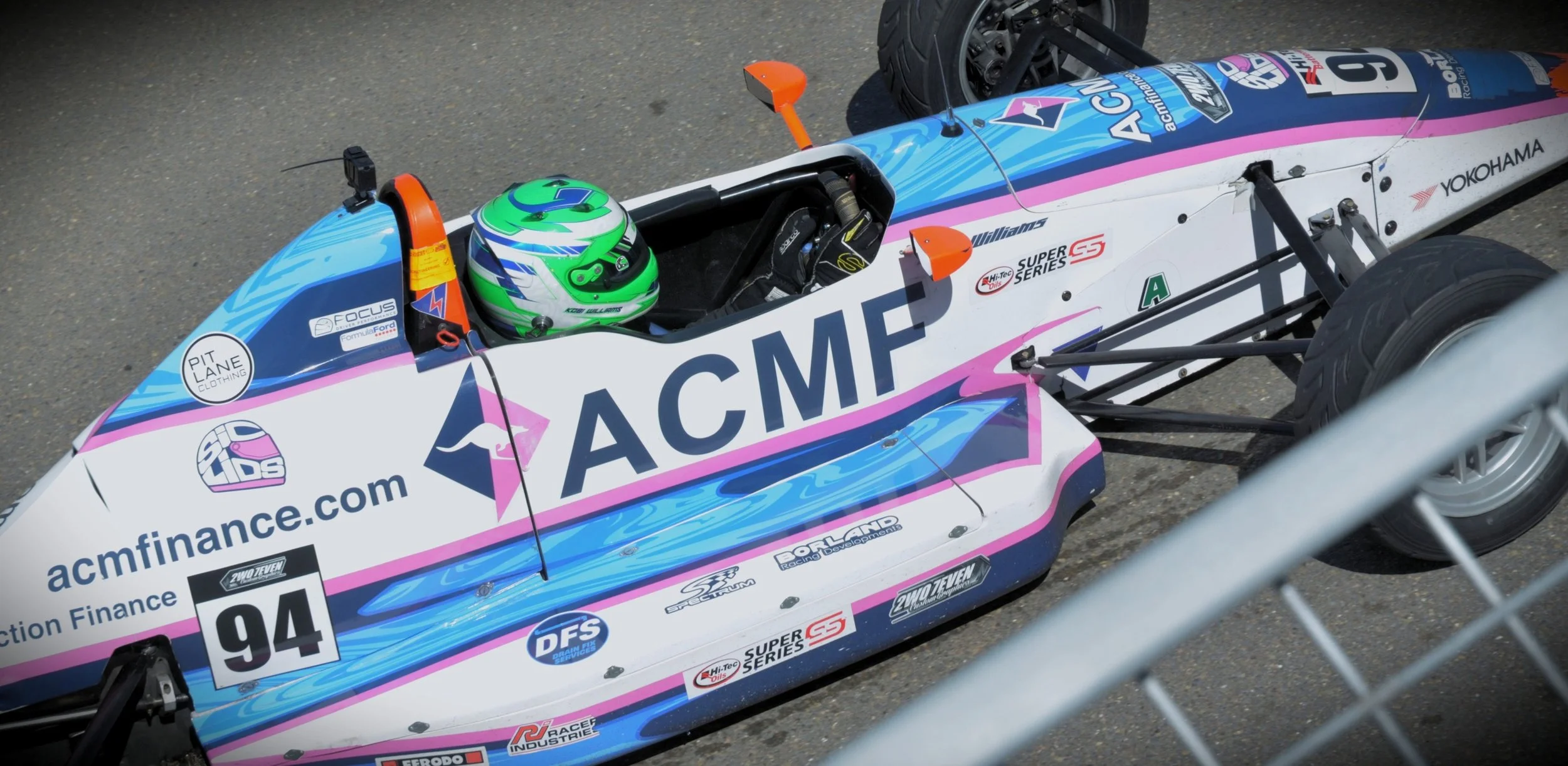 Open cockpit race car with driver in green and white helmet, sponsored by ACME, number 94, on a paved track.