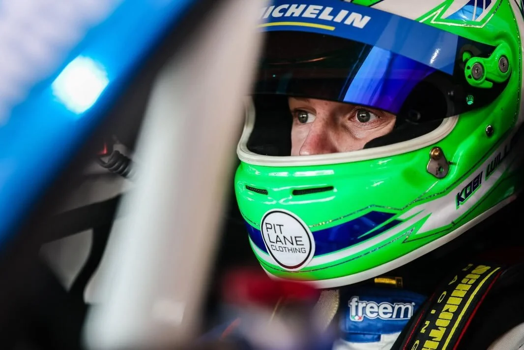 Close-up of a race car driver wearing a green and blue racing helmet, looking focused inside the cockpit.
