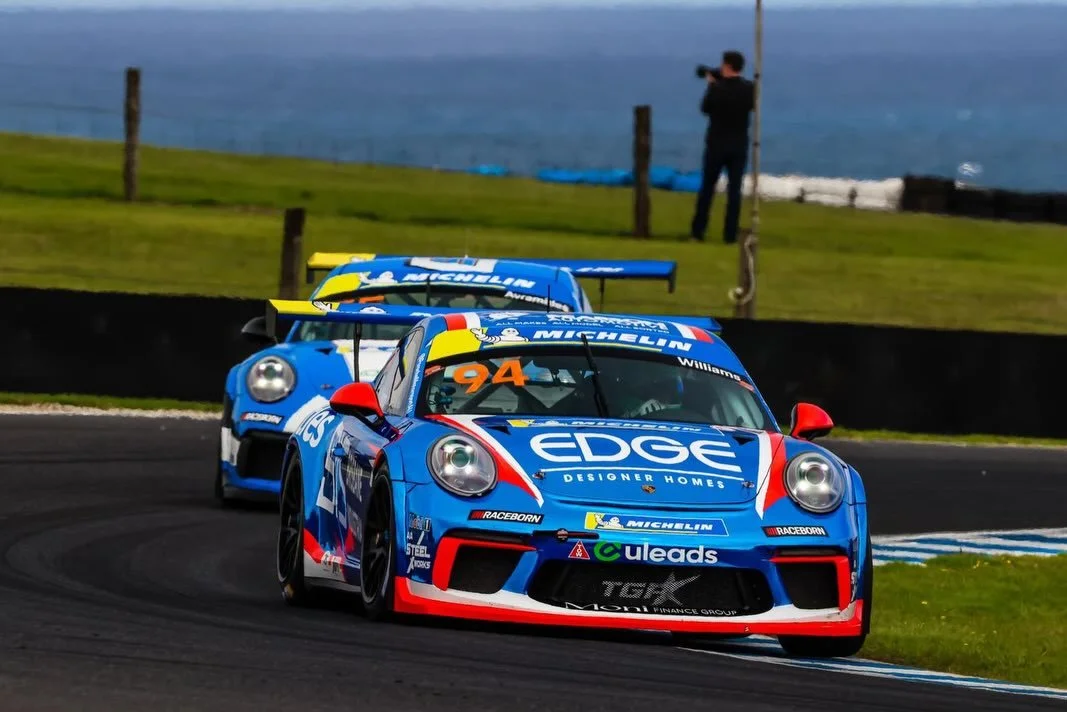 Three racing cars in blue with red and white accents are on a race track, with a person filming them in the background near the ocean.