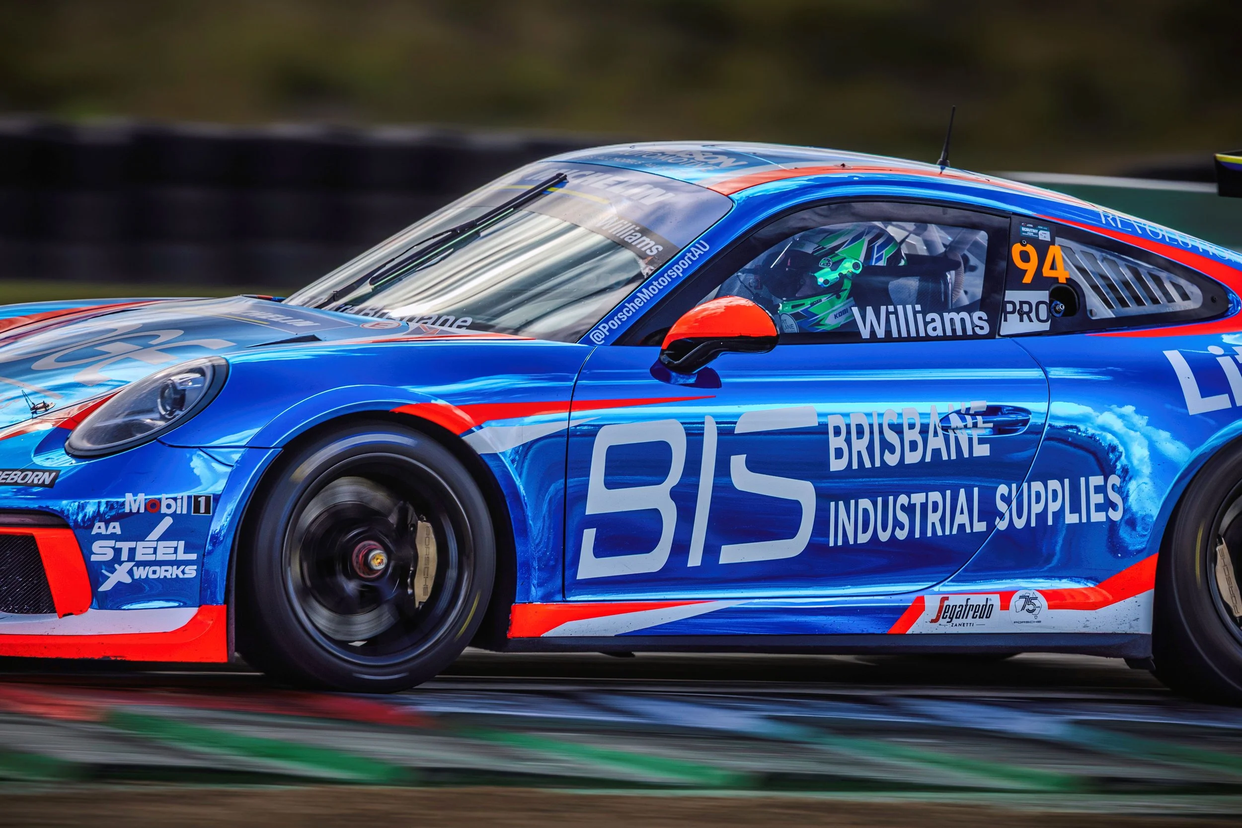 A blue race car with red and white accents speeds along a track, with a driver in a green helmet visible through the window.