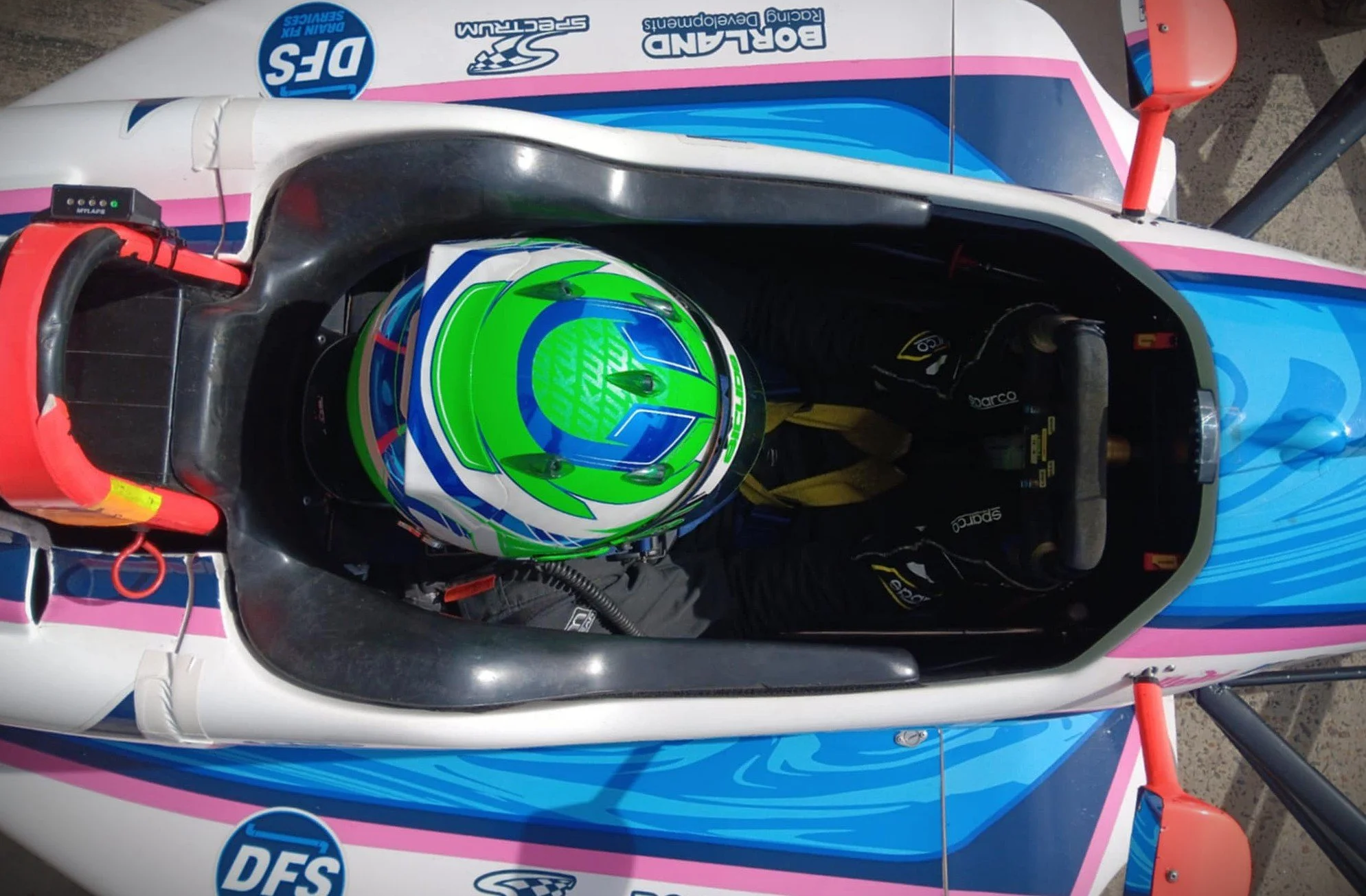 Top-down view of a race car driver in a cockpit, wearing a colorful helmet, with a racing suit and gloves, inside a race car with sponsor logos, including DFS and Bertland.