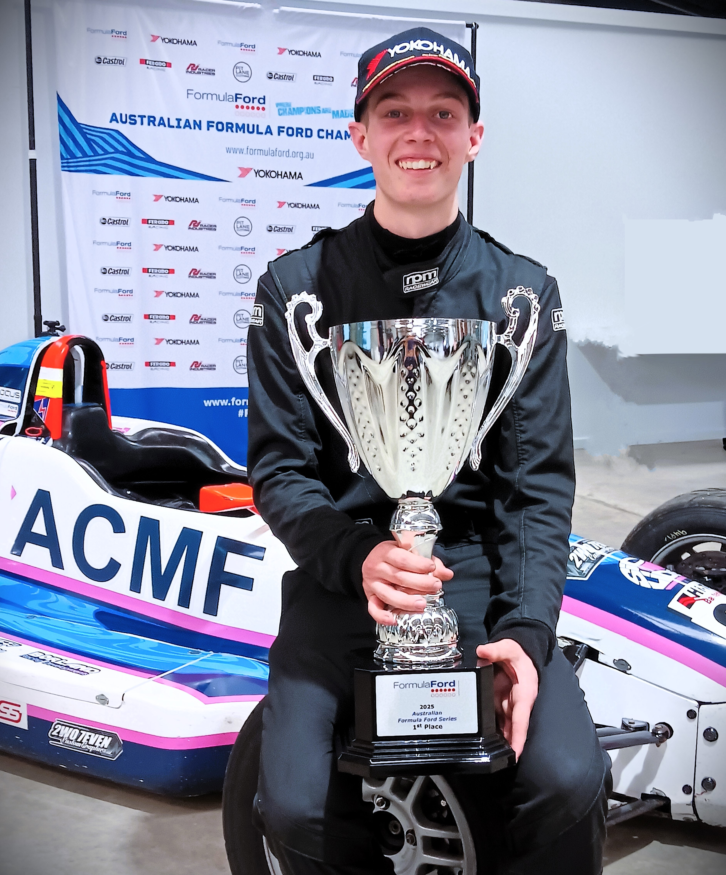 Young male race car driver in a black racing suit holding a silver trophy, sitting on a racing tire, with a race car and a backdrop displaying 'Australian Formula Ford Championship' and sponsor logos behind him.