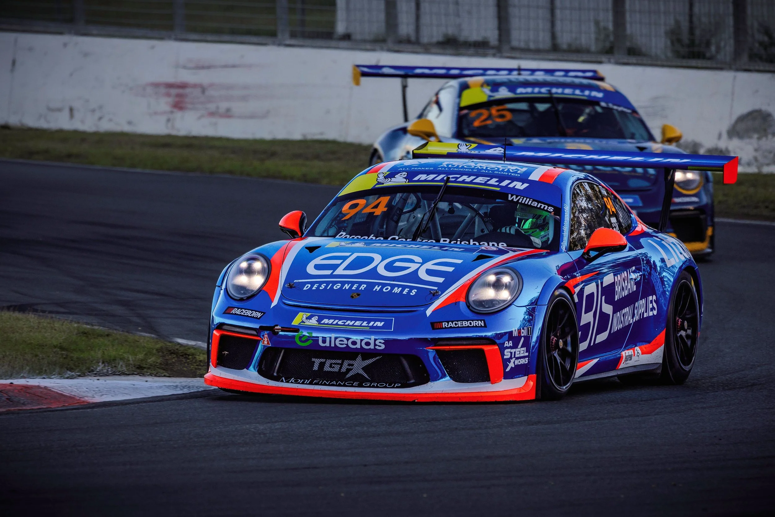 A blue race car with orange accents and sponsor logos, driven by a helmeted driver, is on a race track. Two additional race cars follow behind, also with sponsor branding, as they navigate a turn.
