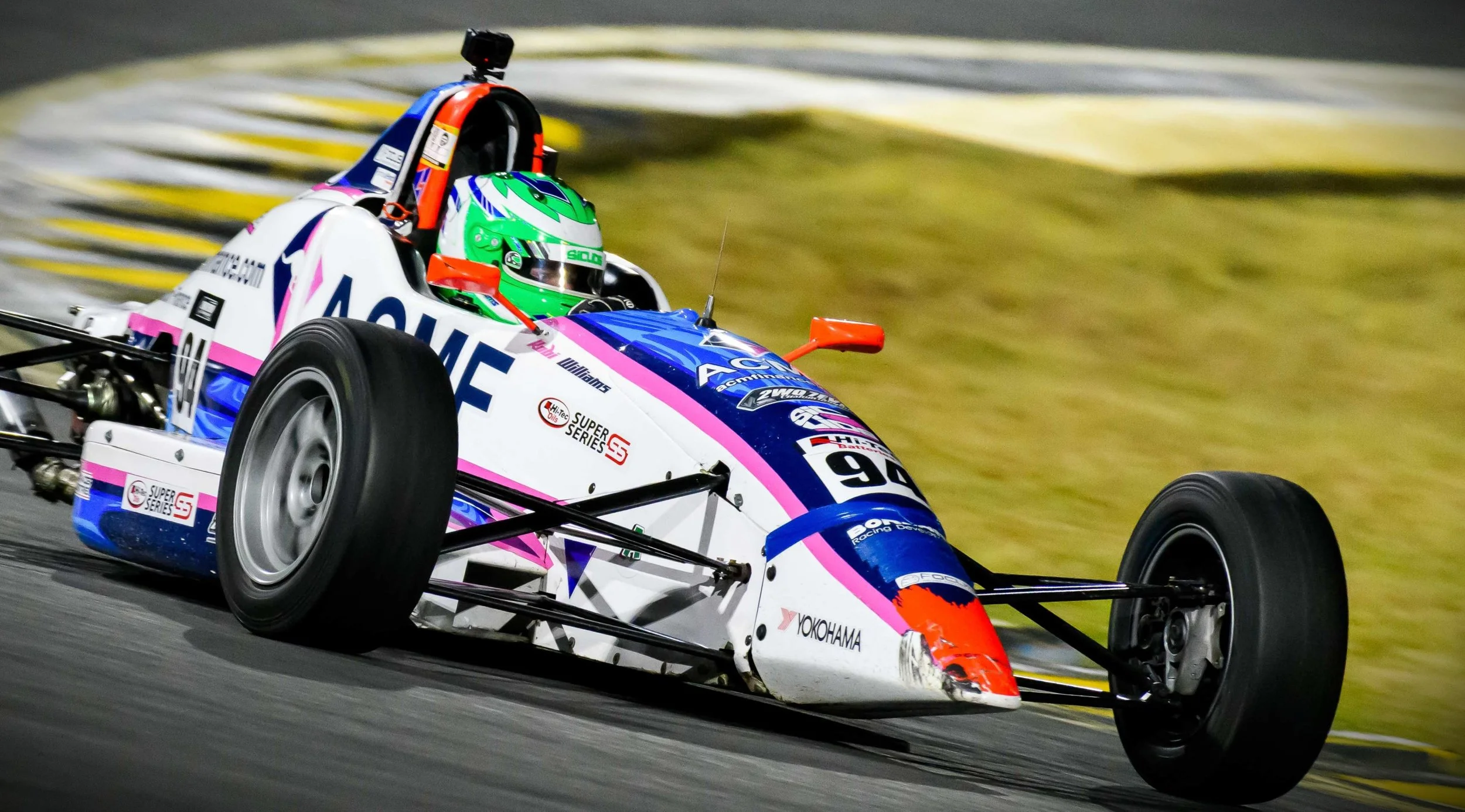 A race car speeding on a track, driven by a driver wearing a green helmet, with a colorful white, pink, and blue livery decorated with sponsorship logos.