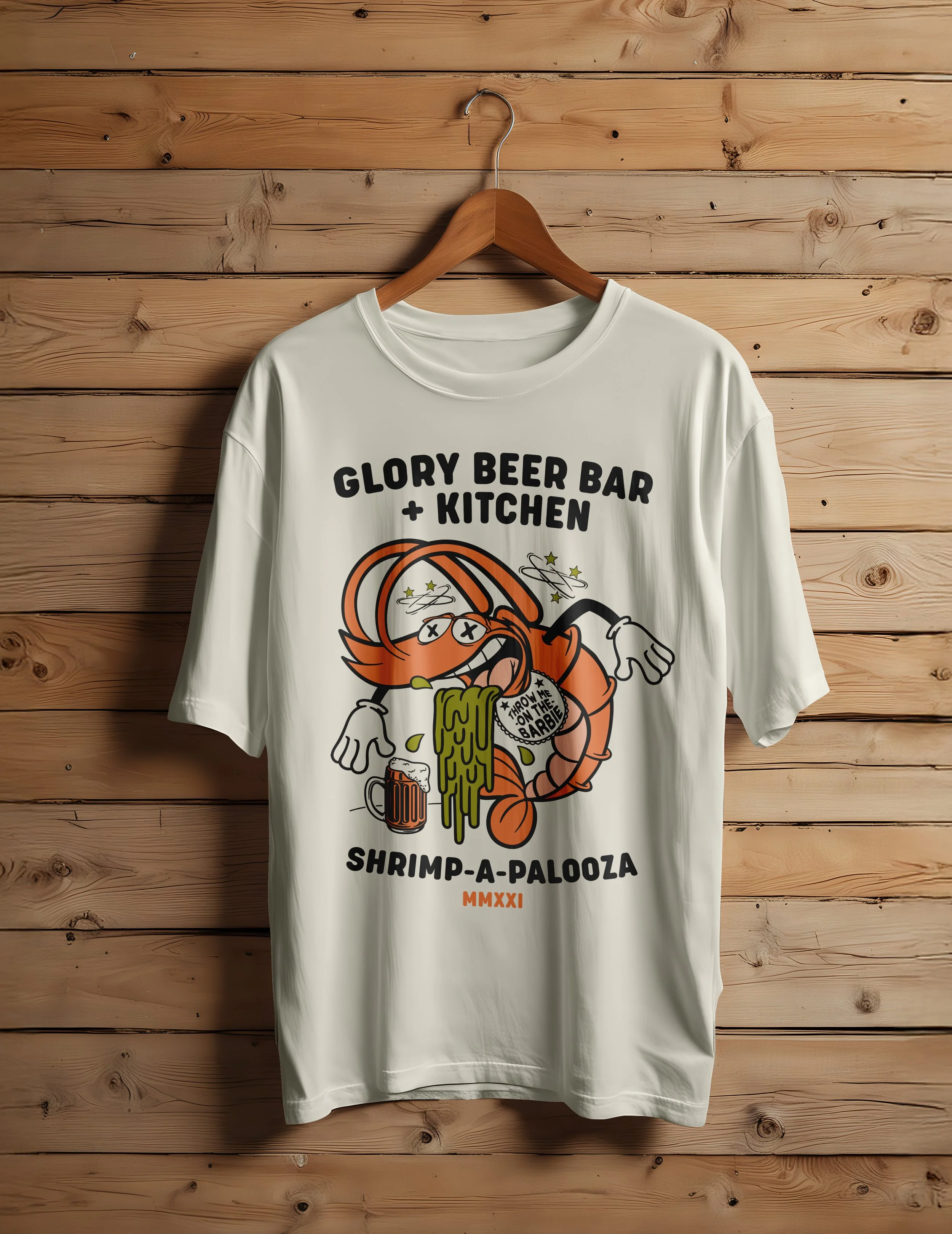 White T-shirt hanging on a wooden wall, featuring a cartoon lobster holding a beer mug with the text 'Glory Beer Bar + Kitchen Shrimp-A-Palooza MMXXI'.