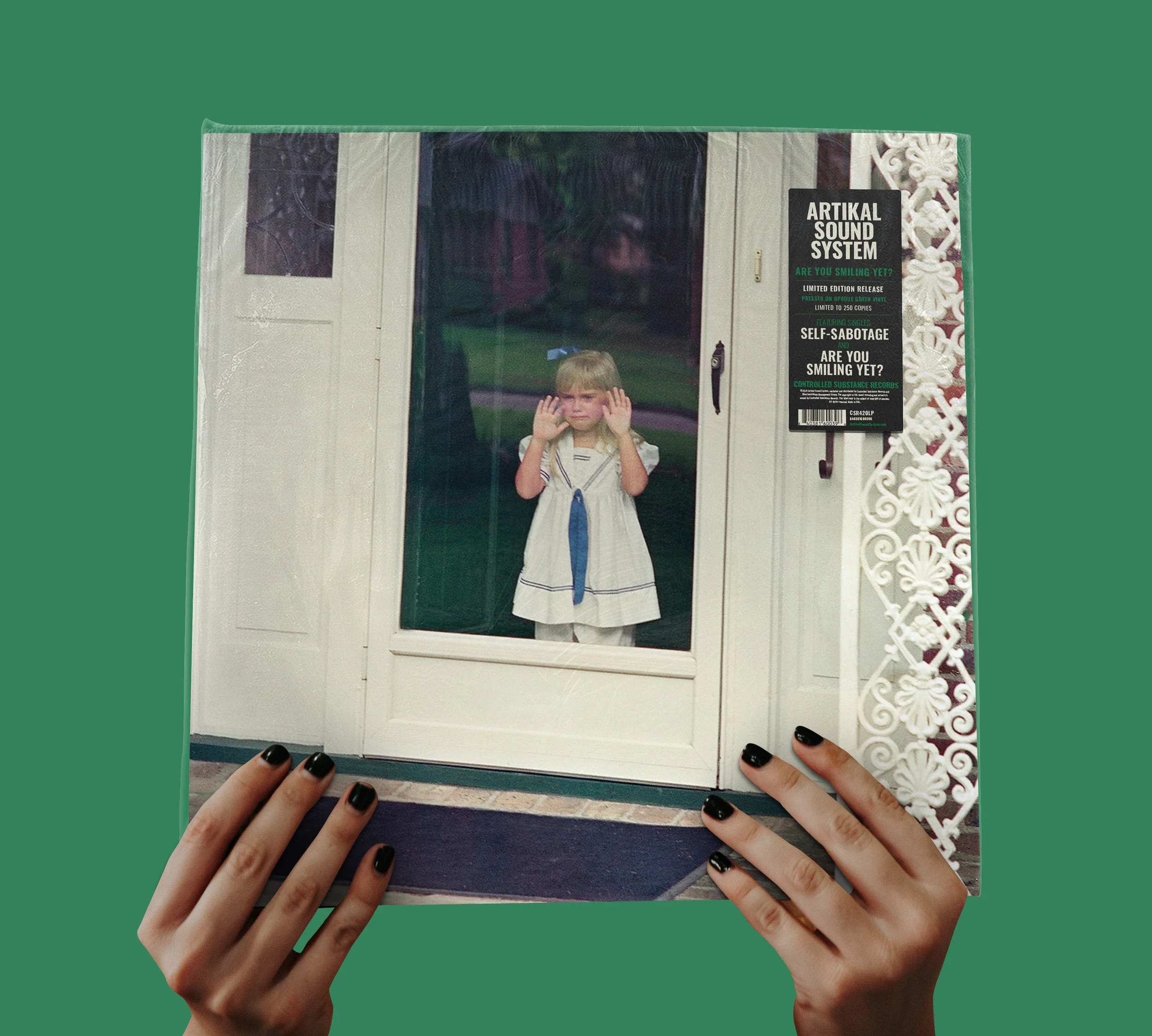 A person holding a framed picture of a young girl looking out a front door glass window with her hands on the glass. The girl is wearing a dress with a sailor collar and a blue ribbon, and the scene is outdoors with a green lawn.