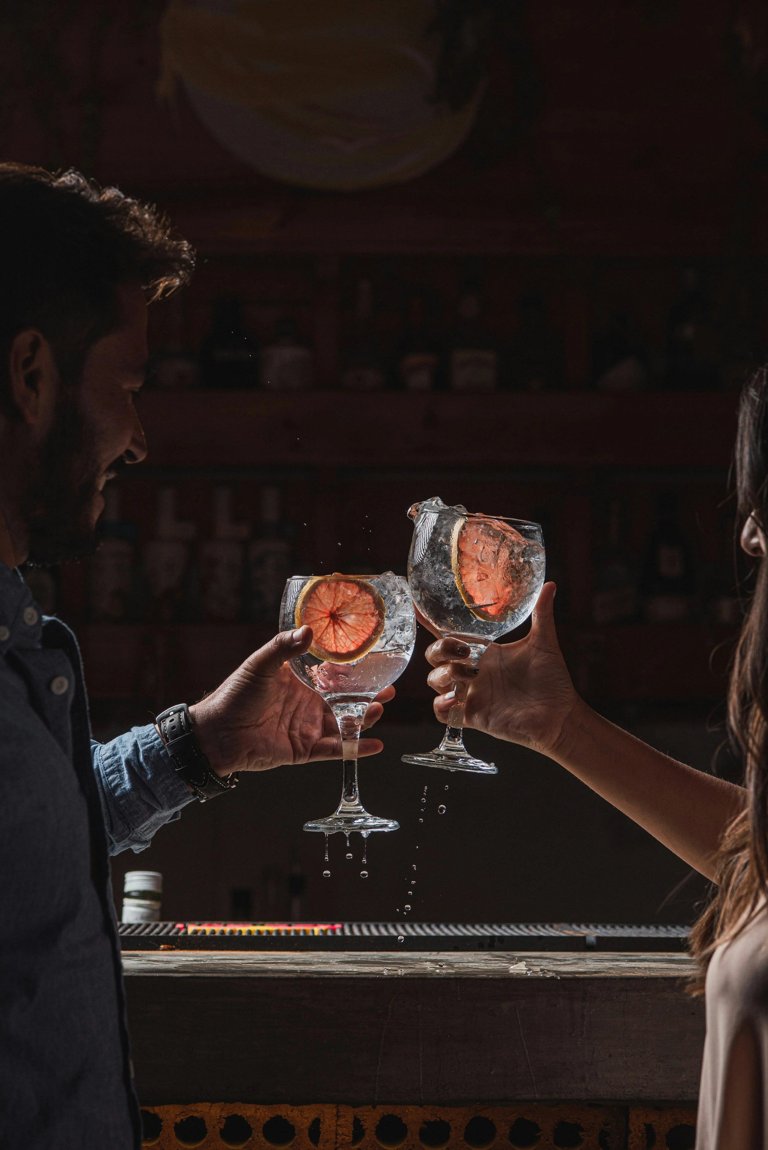 Two people clinking glasses with grapefruit slices inside, water splashing, in a dimly lit bar.