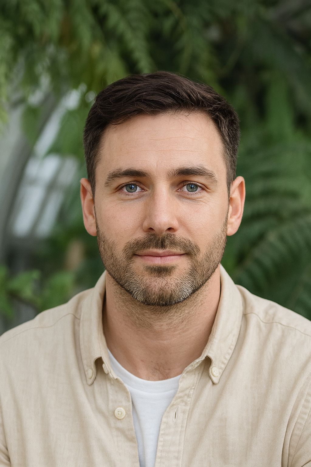 Close-up of a man with short dark hair, blue eyes, and a beard, wearing a beige shirt, in front of green foliage.