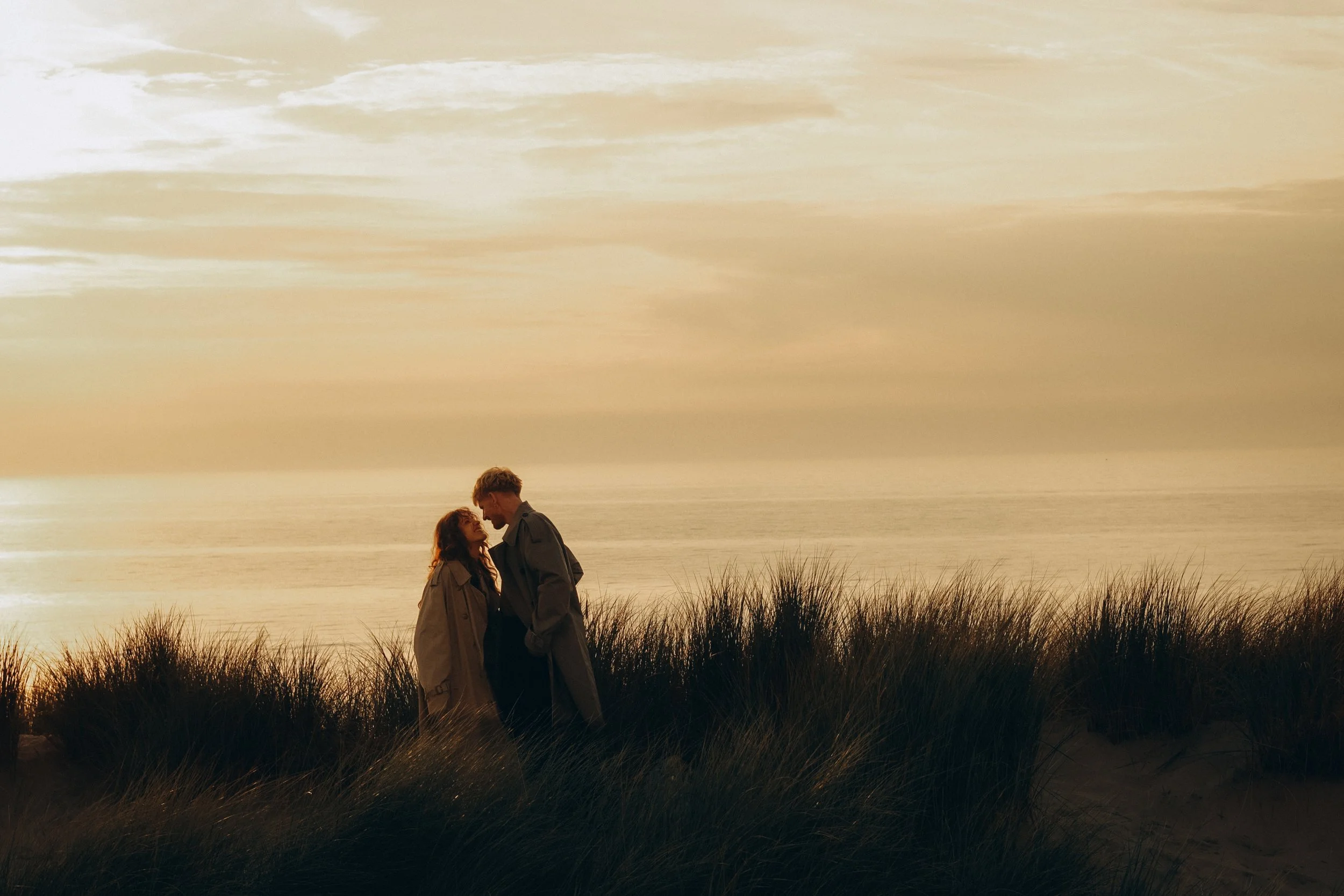 A couple stands close together on a sandy beach with grass in the foreground, facing each other under a sunset sky.