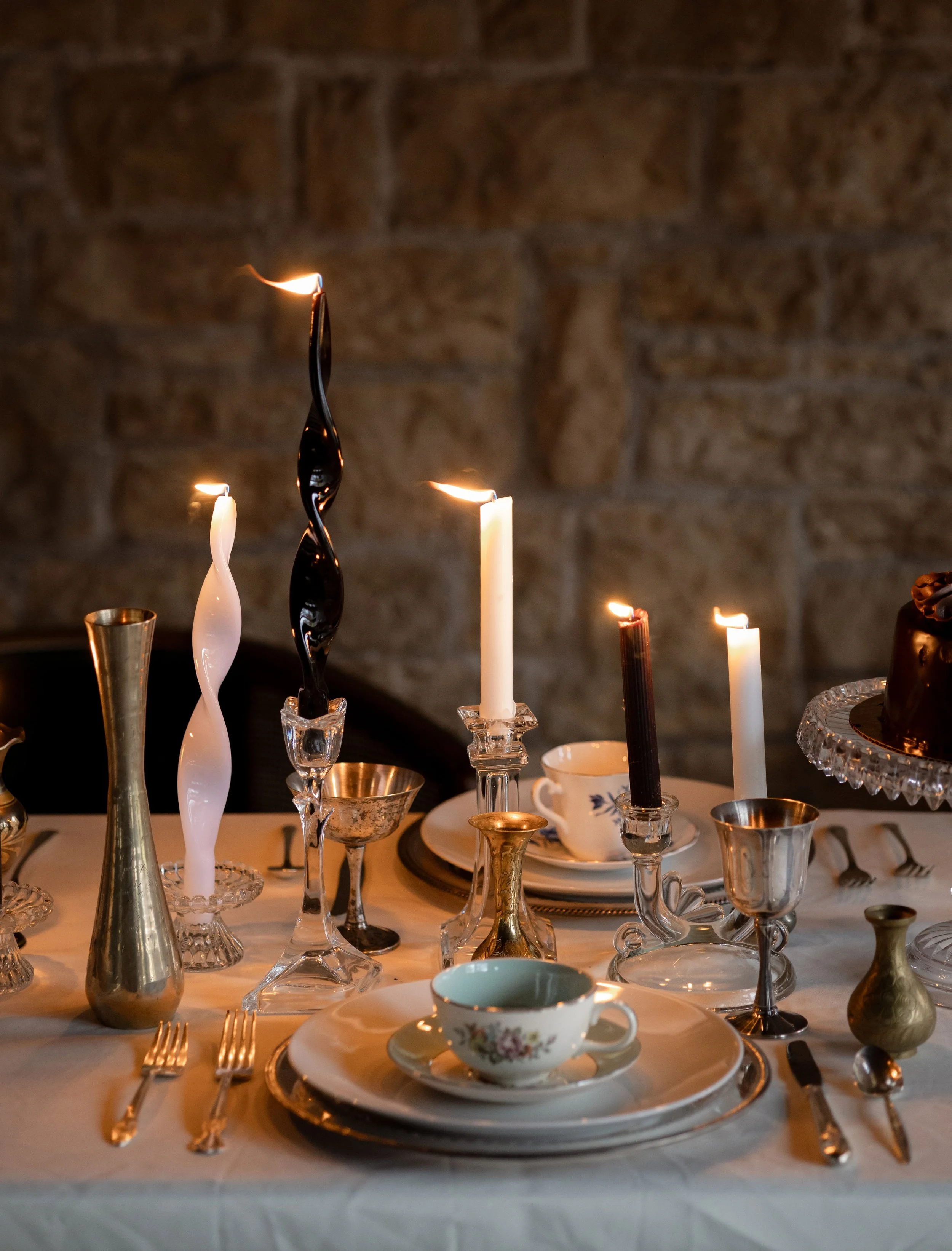 An elegant table setting with various glassware, candles, and fine china on a white tablecloth, against a brick wall background.