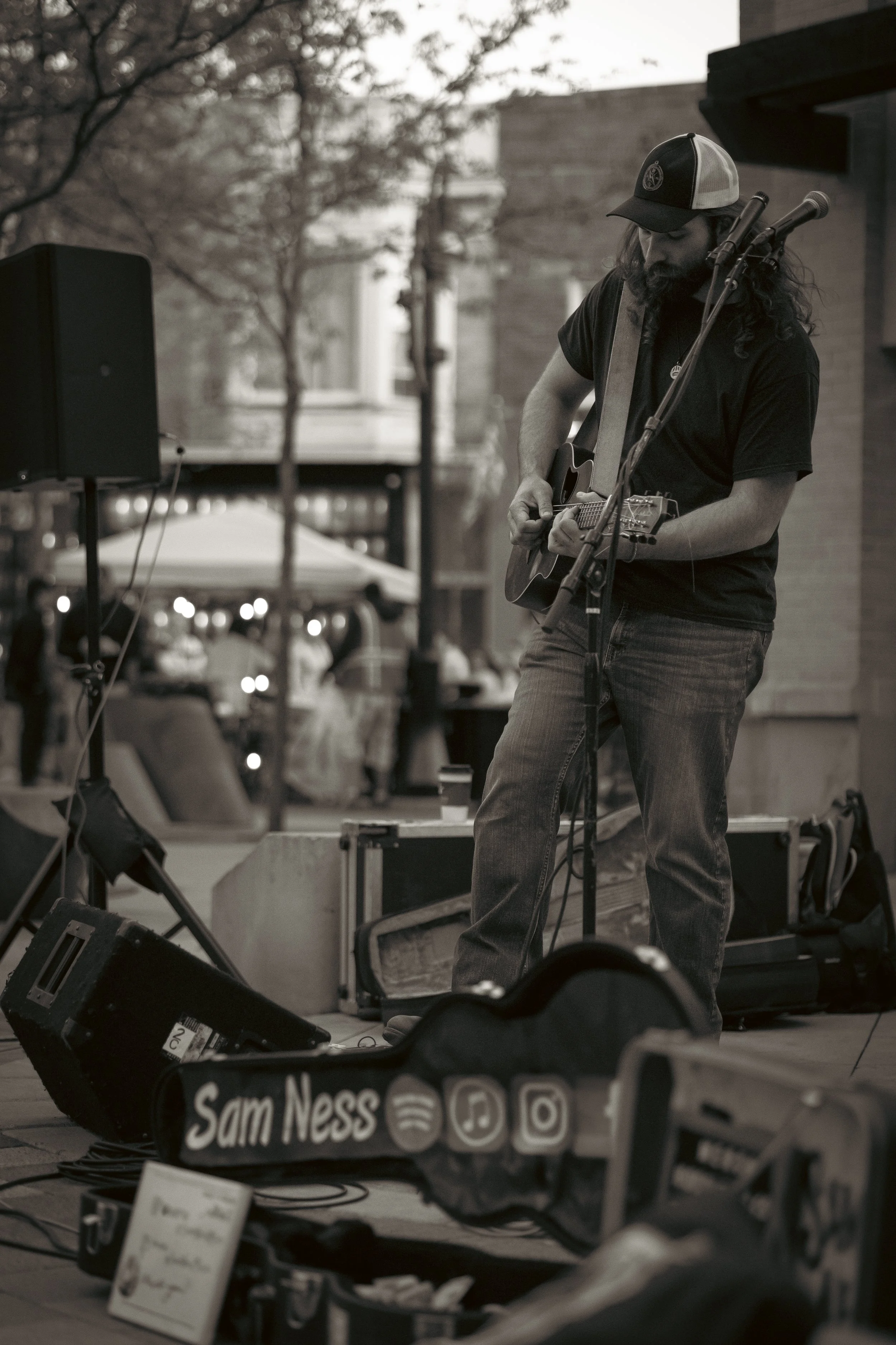 A man with long hair, beard, and a cap is playing an acoustic guitar on a street during daytime. There is musical equipment and a sign with social media handles in the foreground.