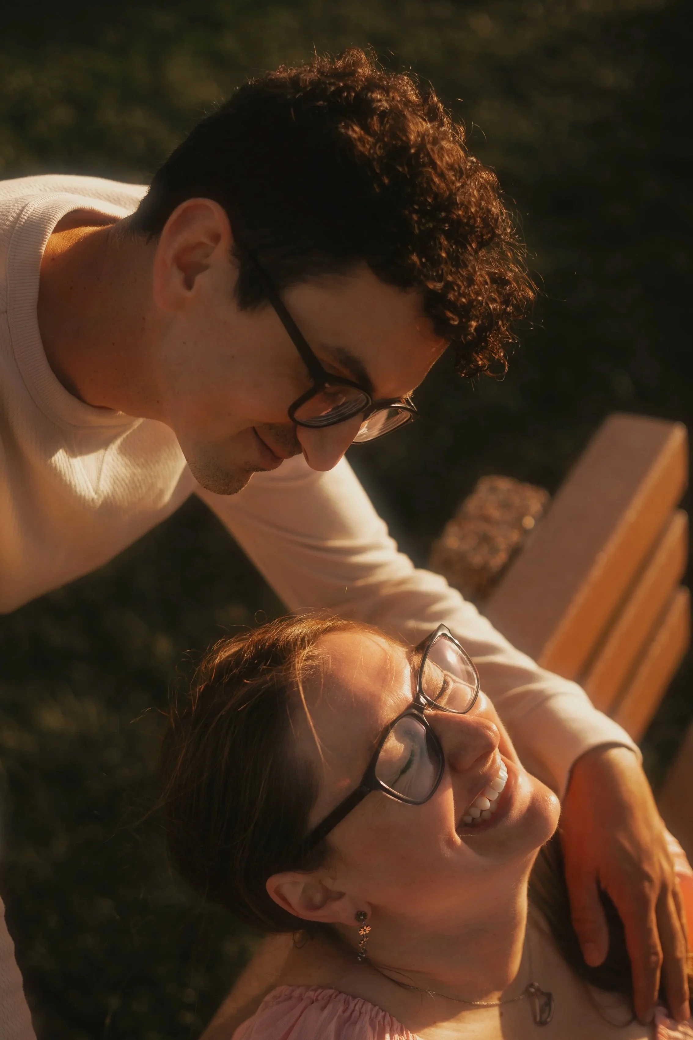 A man and a woman lying outdoors at sunset, both smiling and looking at each other, with the man resting his head next to the woman's head.