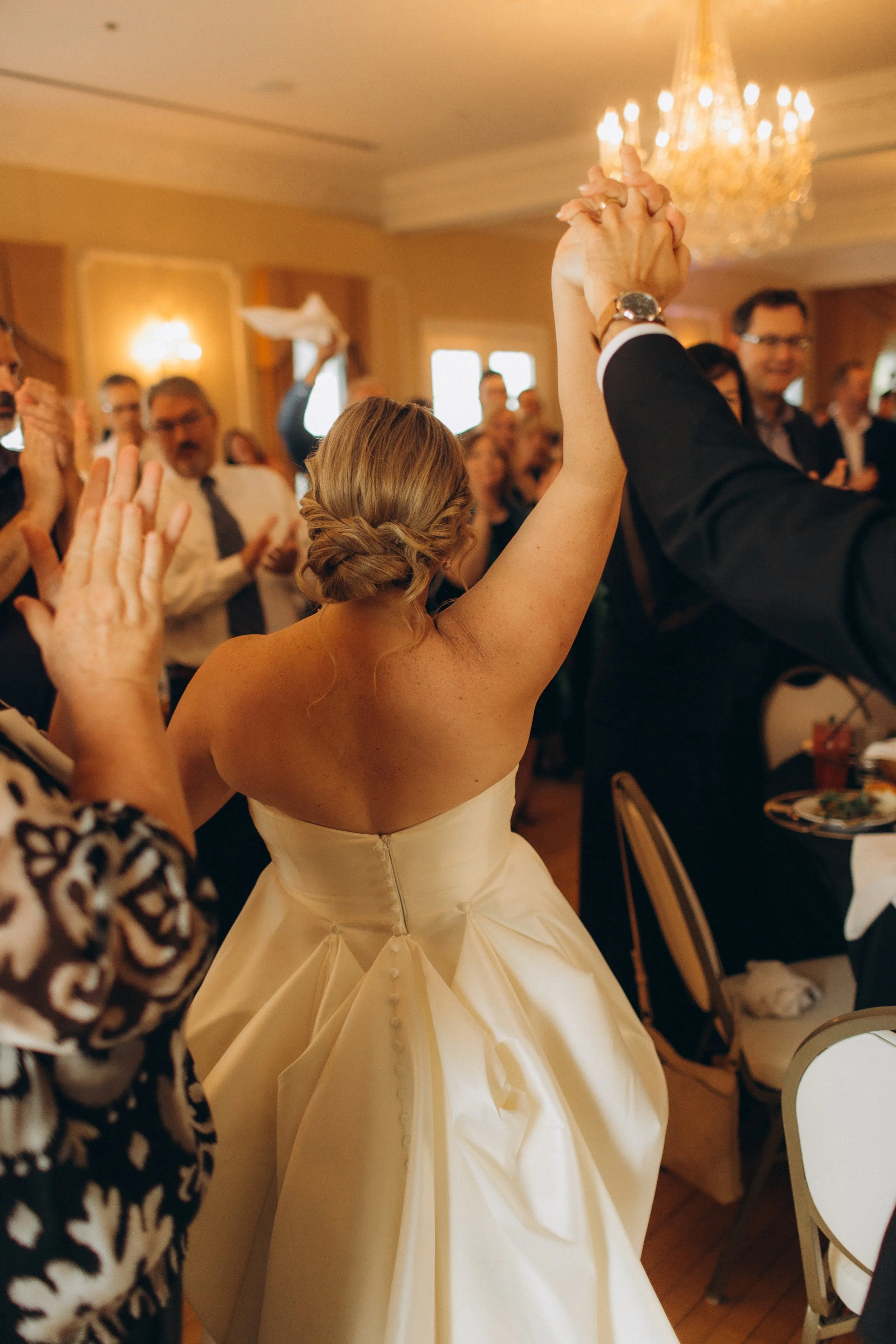 A woman in an elegant wedding dress dancing with a man at a wedding reception. She is holding hands with someone and facing away from the camera. There are guests and a chandelier in the background.