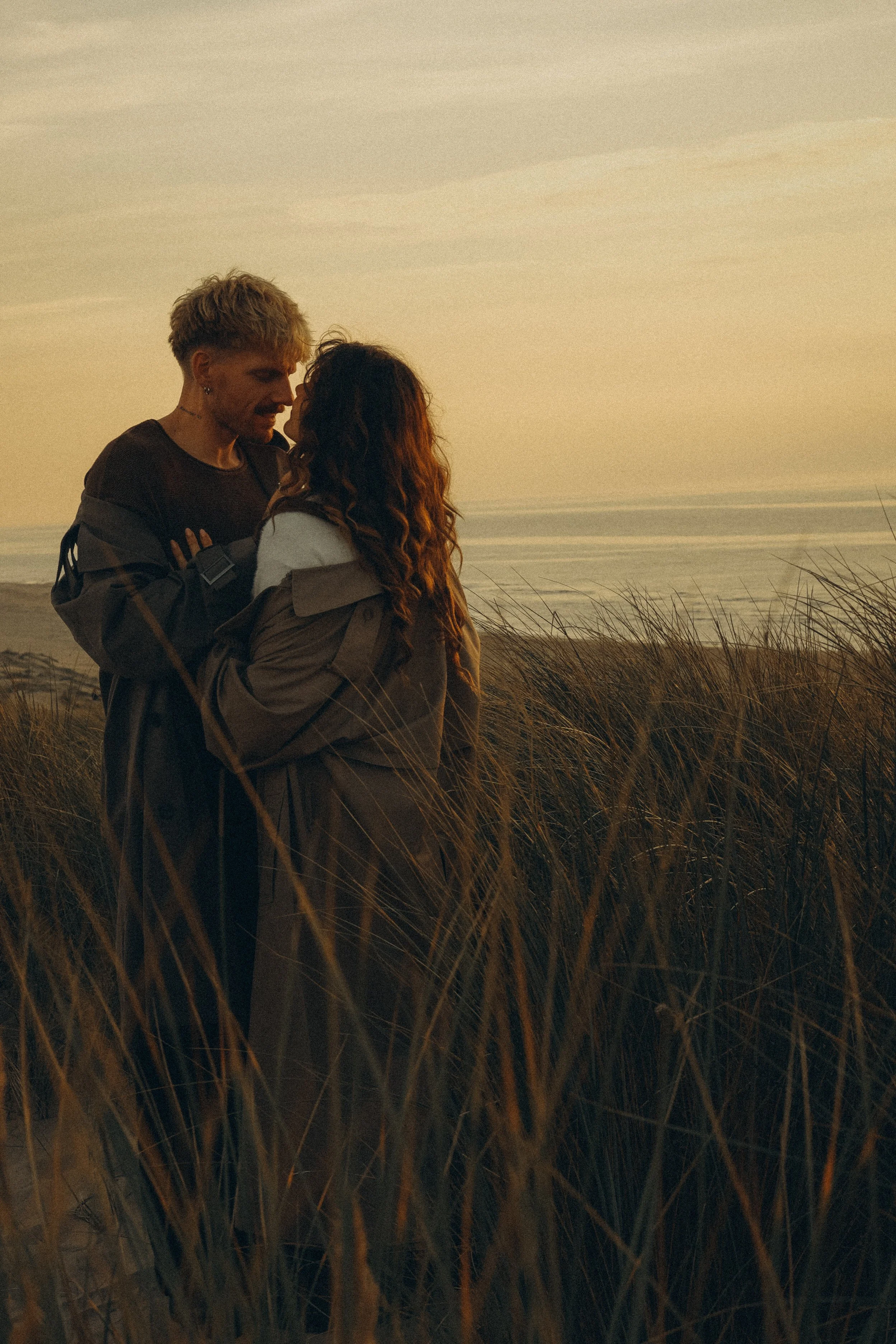 A young couple standing close together on a sandy beach during sunset, surrounded by tall grass and ocean waves in the background.