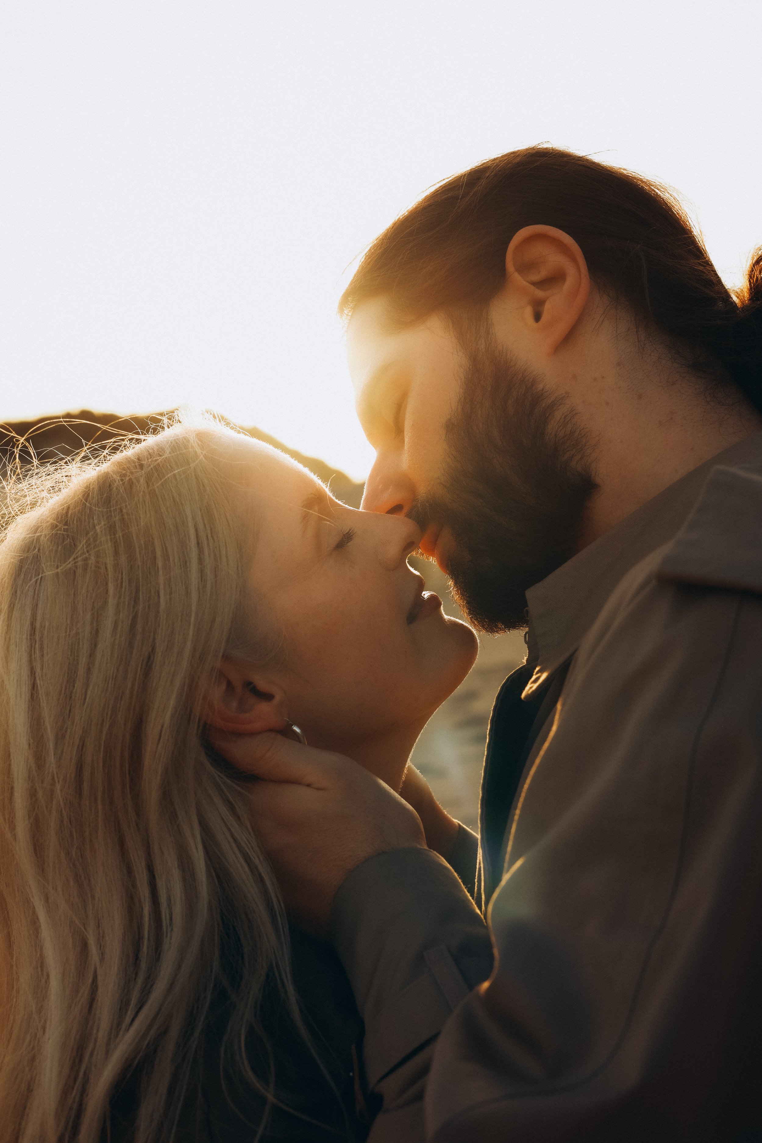 A man and a woman are about to kiss each other in close proximity, with the woman gently touching the man's neck, during sunset or sunrise.