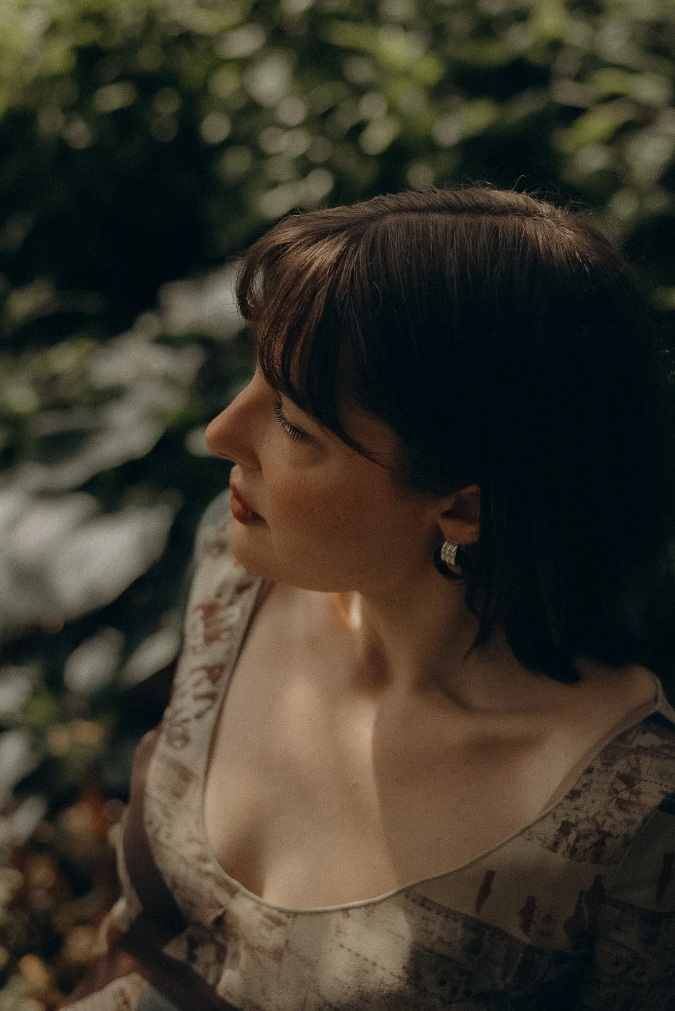 A woman with short brown hair wears a beige and brown patterned dress and pearl earrings. She is sitting outdoors near a stone wall covered with green foliage, looking downward with eyes closed.