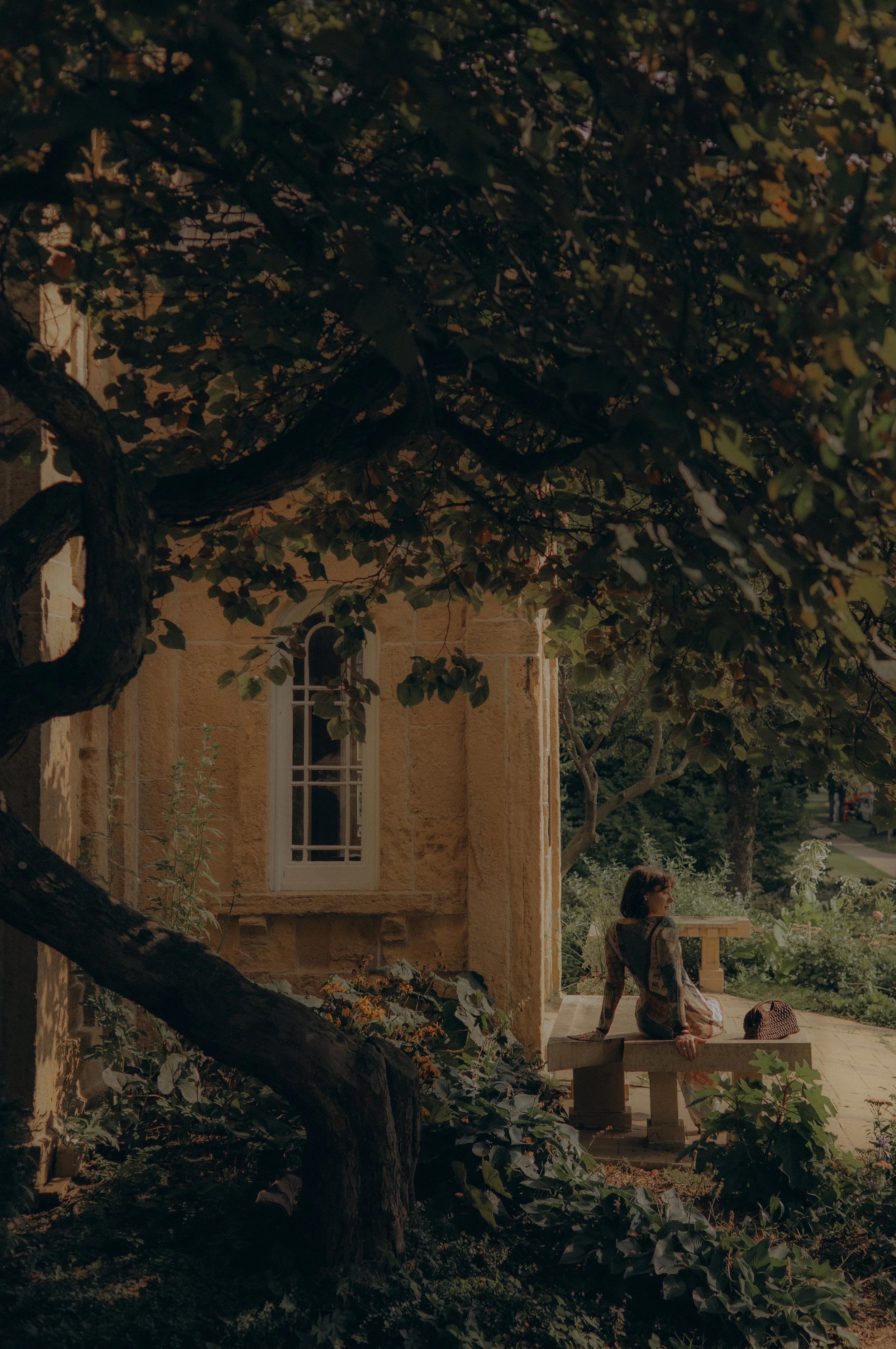 A woman sitting on a stone bench near a tree in front of a beige stone building with arched windows, surrounded by greenery, during daytime.