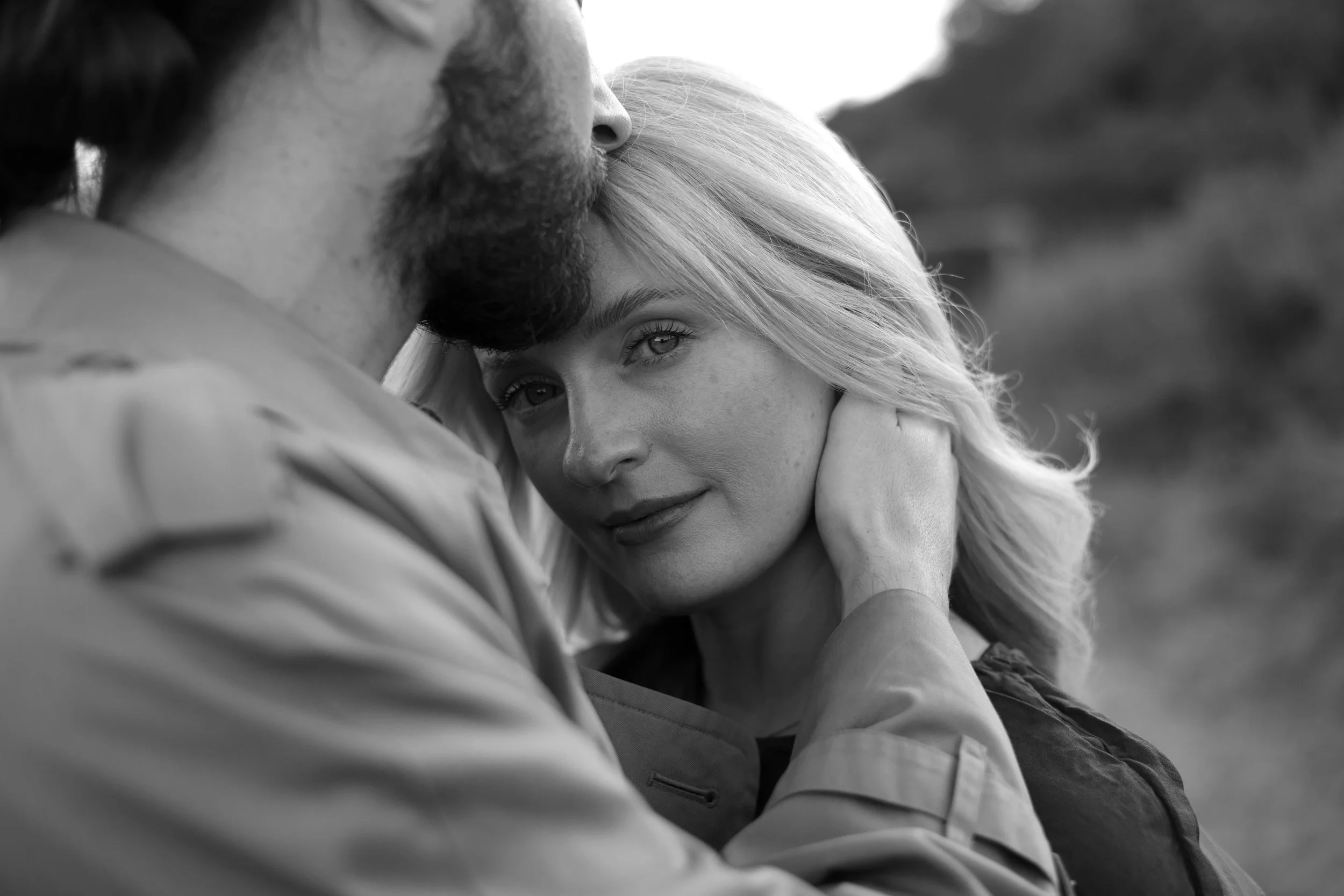 A black and white close-up photo of a couple in an intimate moment, with the woman resting her head on the man's shoulder, both smiling softly.