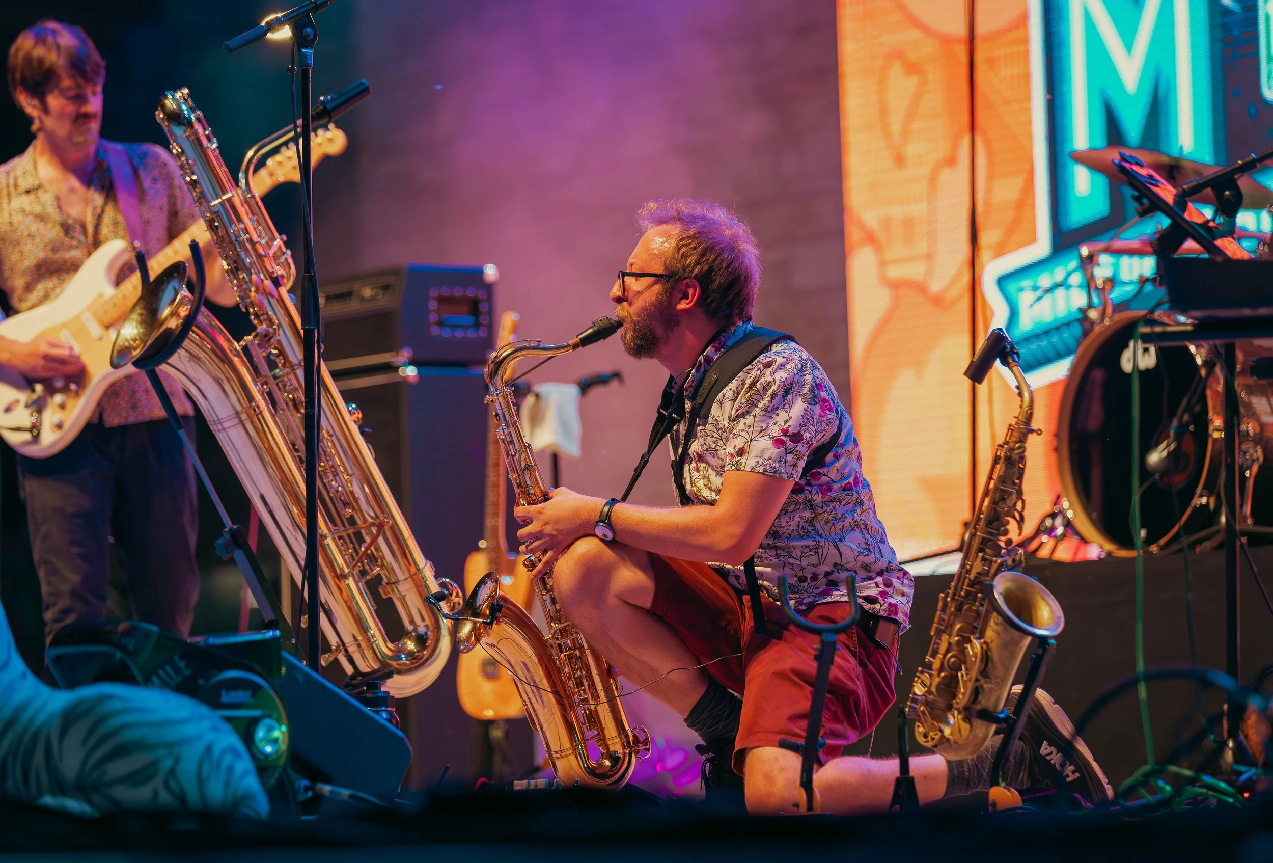 Musicians performing on stage with saxophones, electric guitar, and keyboards, illuminated by colorful stage lighting and neon signs.