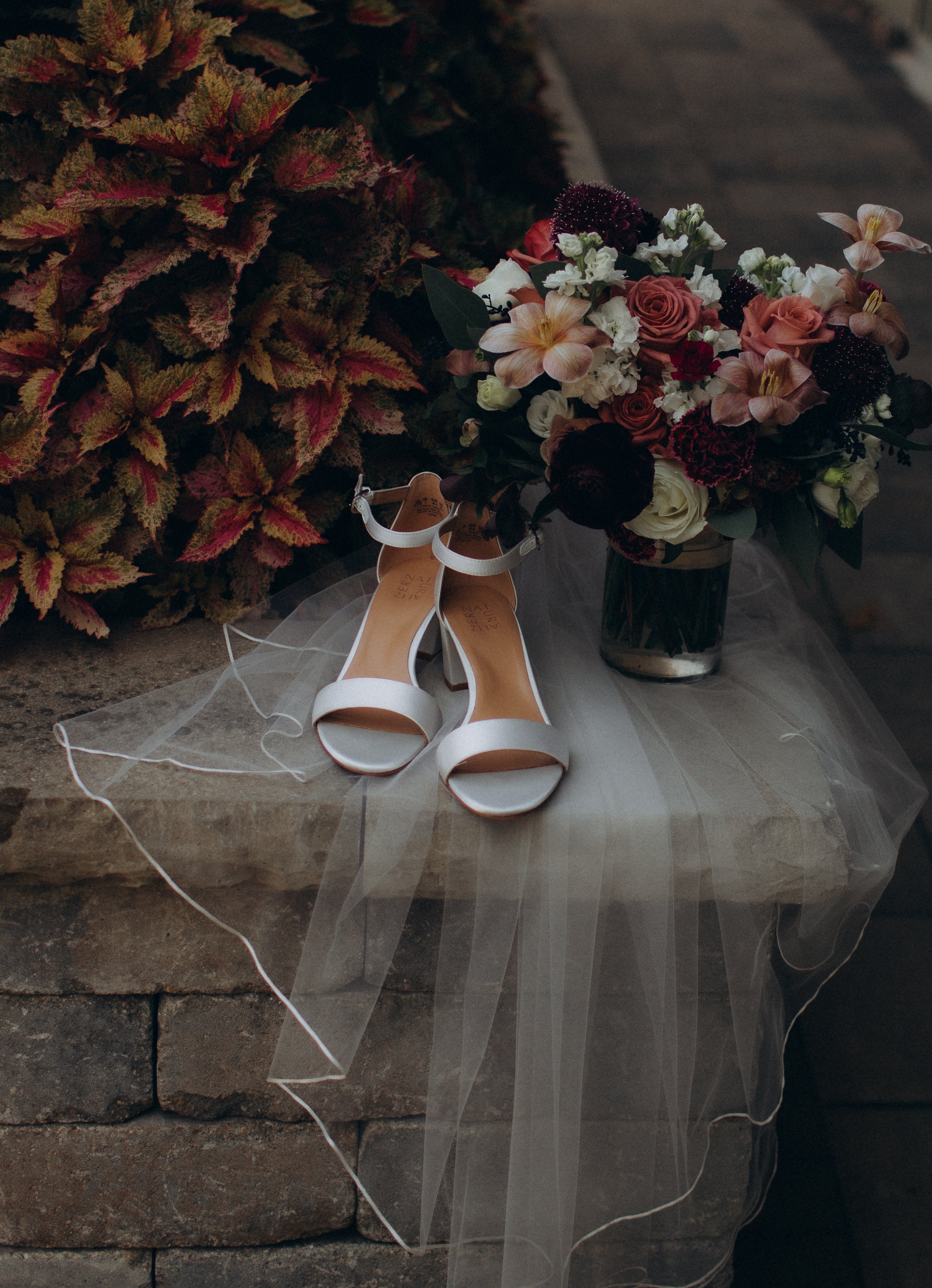A pair of white high-heeled wedding shoes with ankle straps placed on a rock surface next to a large bouquet of mixed flowers including roses, orchids, and other blooms, with a sheer veil spread out on the ground.