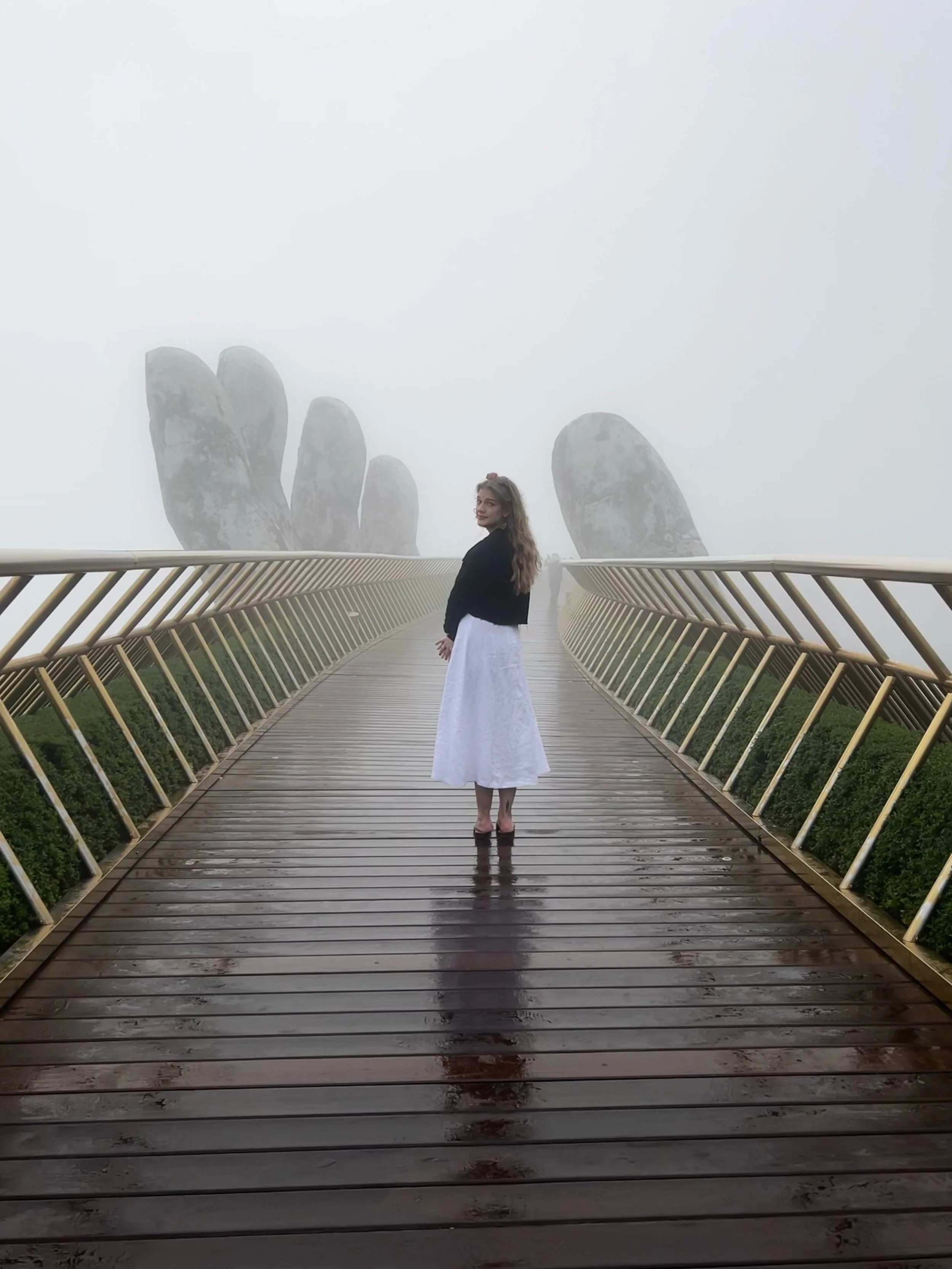 A woman in a black jacket and white skirt stands on a wooden bridge in front of large fog-shrouded rock formations, with some green bushes visible at the sides of the bridge.