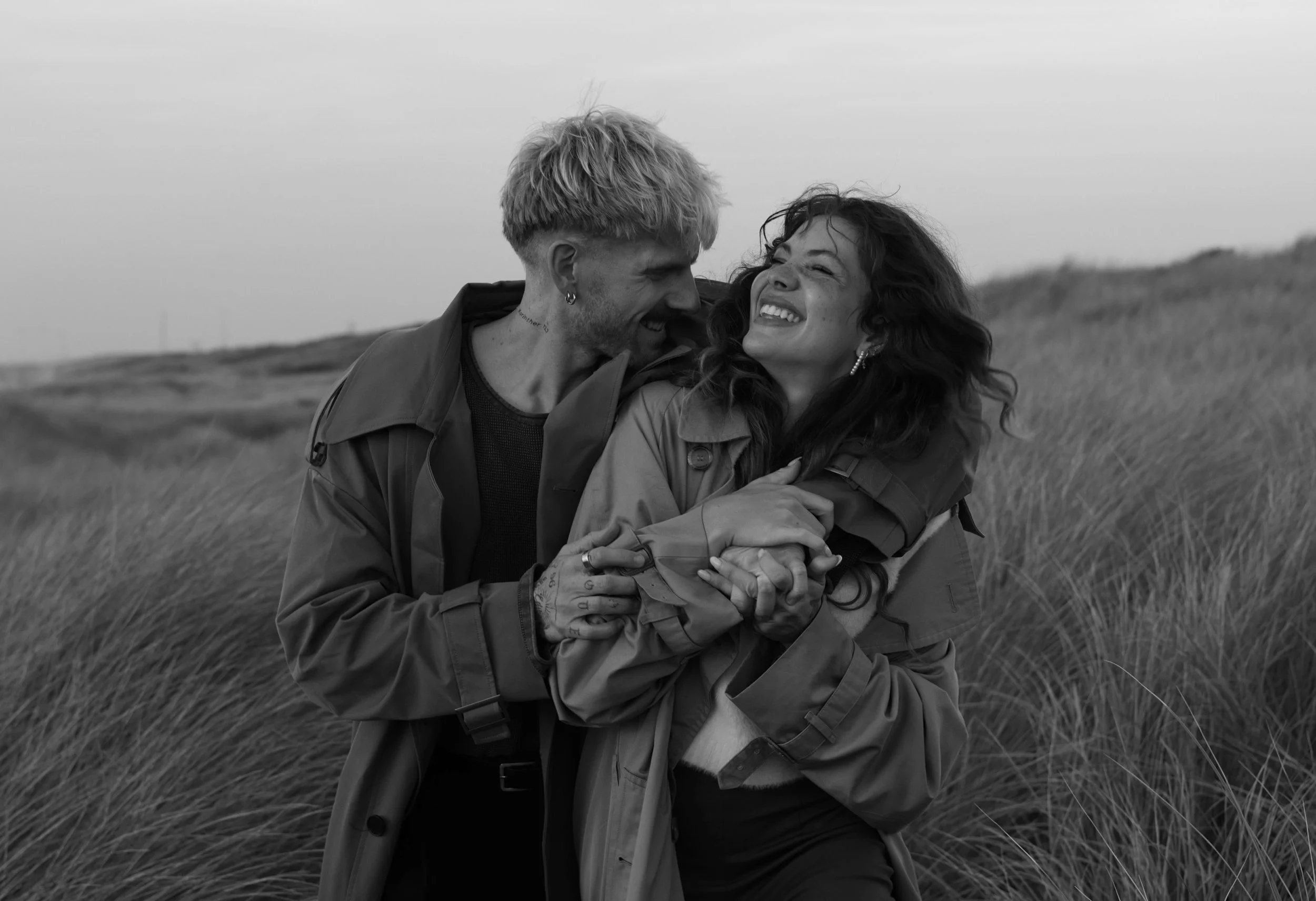 A black and white photograph of a couple outdoors in a grassy field, smiling and embracing each other.