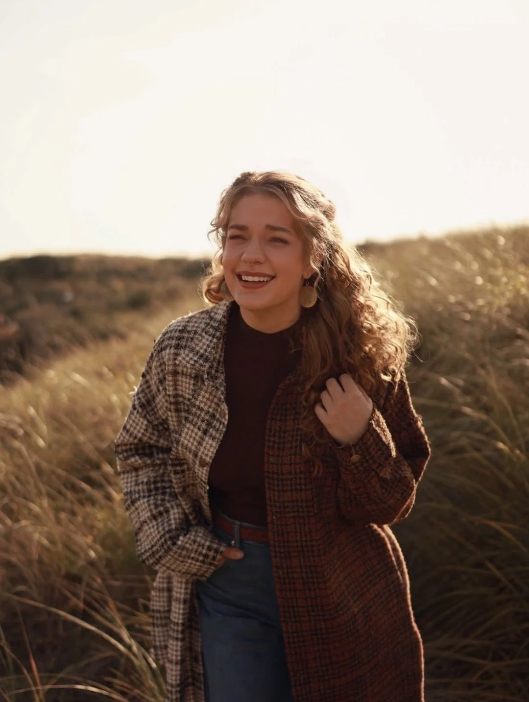 A woman with curly hair smiling in a field of tall grass during sunset, wearing a plaid coat over a dark top and jeans.