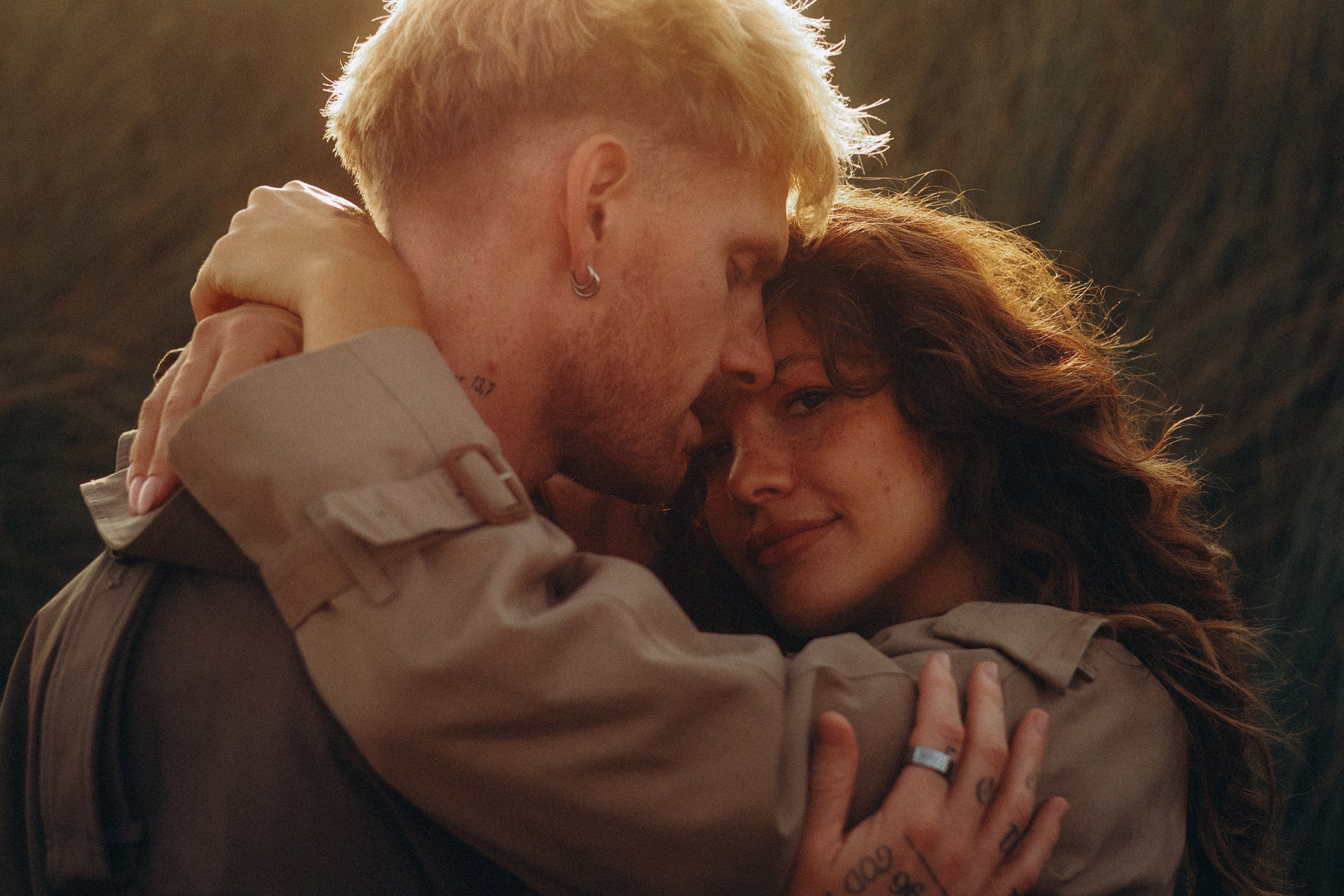 Two people are hugging closely, with their faces touching. One person has short blond hair, an earring, and tattoos, while the other person has long, wavy auburn hair and tattoos on their hand. They are embracing each other tenderly.