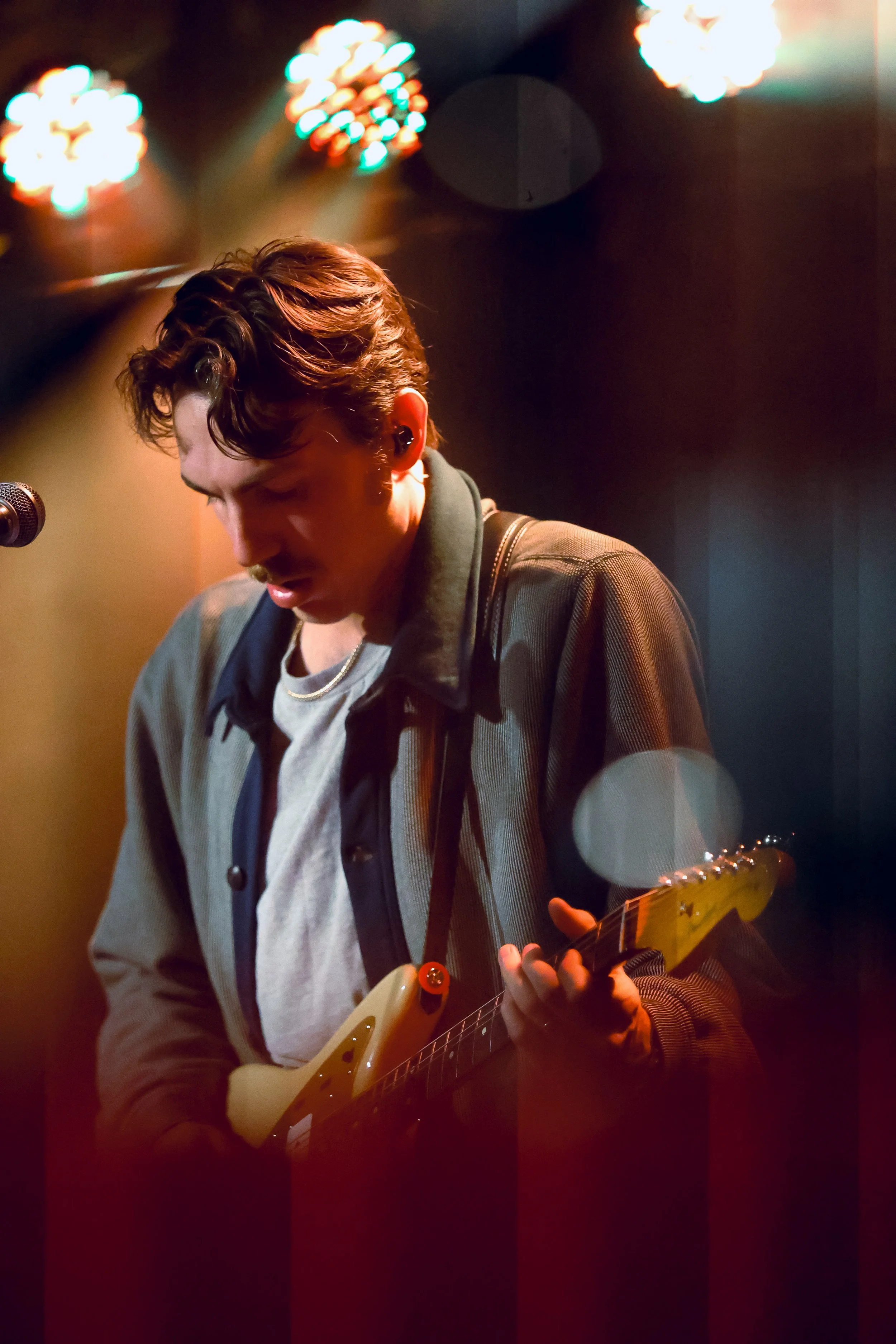 A young man with dark wavy hair playing an electric guitar on stage under colorful stage lights, wearing a gray t-shirt, a jacket, and a necklace.