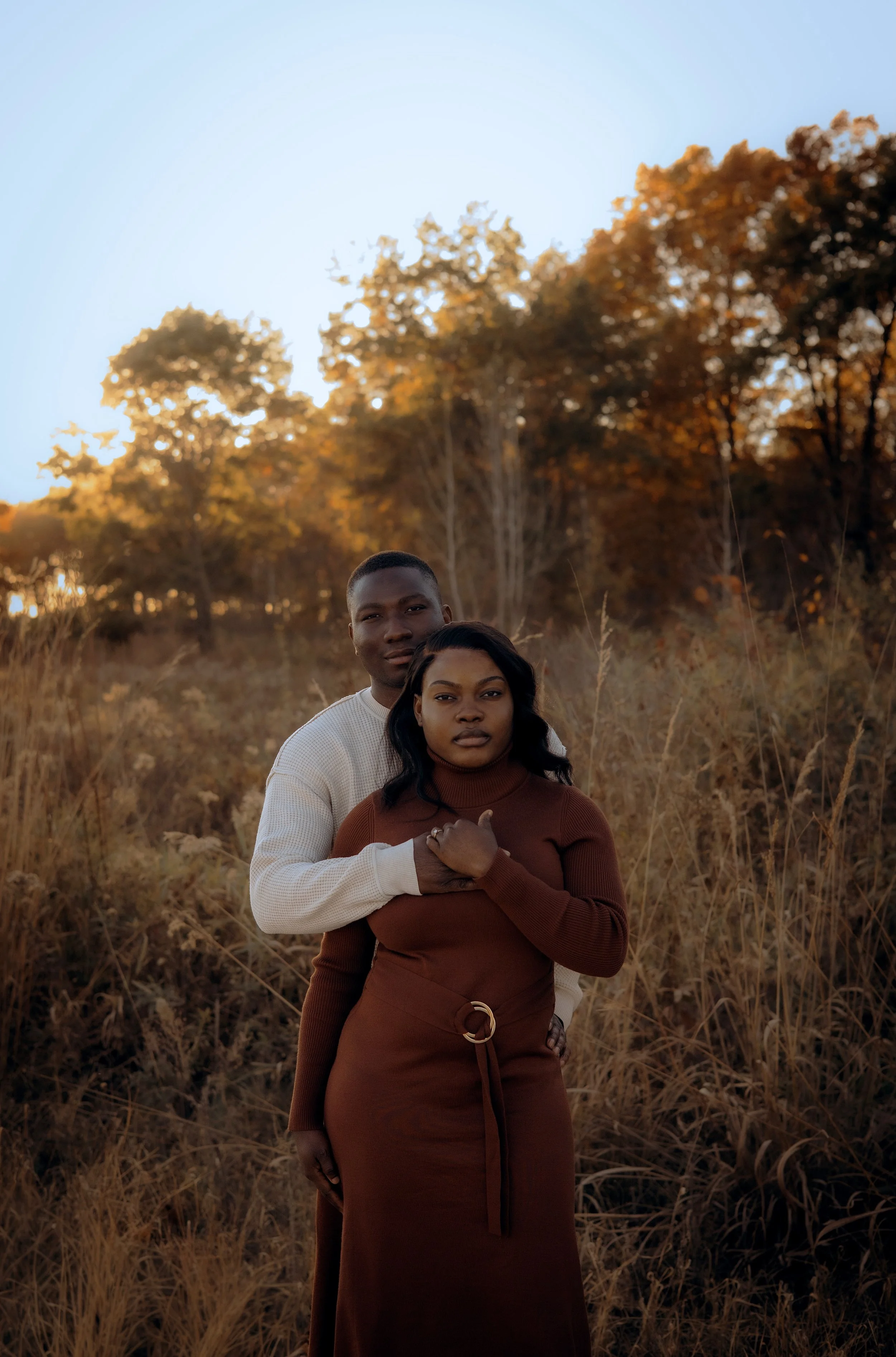 A young couple standing outdoors in a natural setting during autumn, with trees displaying fall foliage in the background. The woman is in front, wearing a brown dress, and the man is standing behind her with his arms around her waist, both looking a