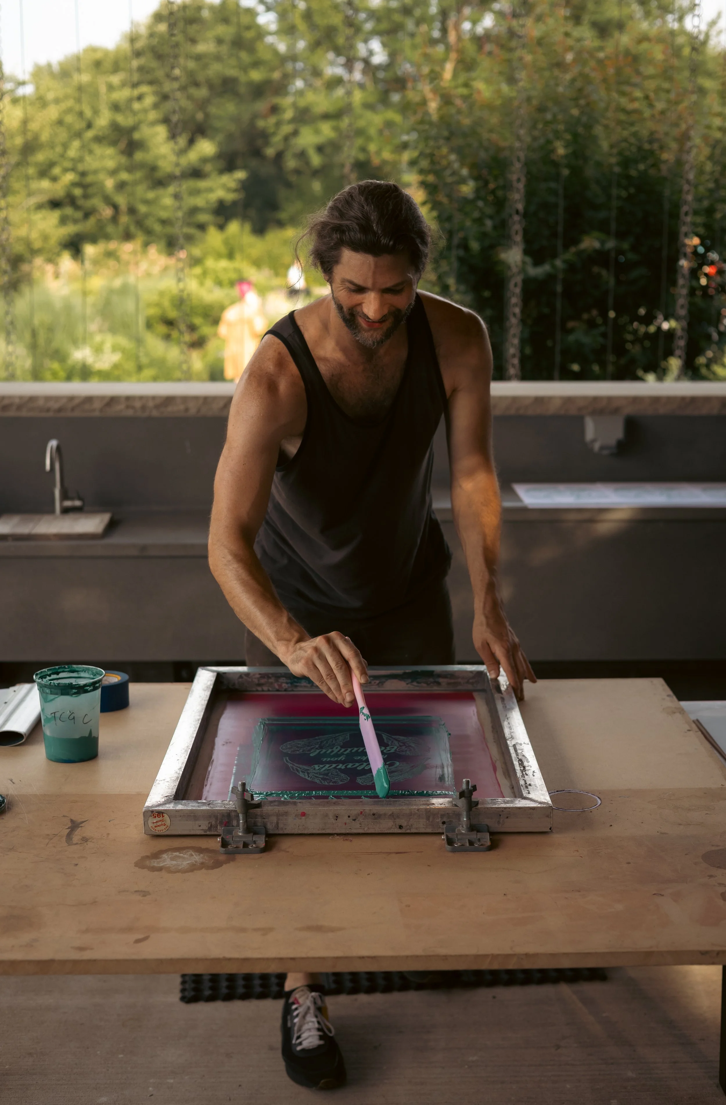 A man in a black tank top and sneakers, smiling and working on a screen printing project at a table, with a nature view in the background.