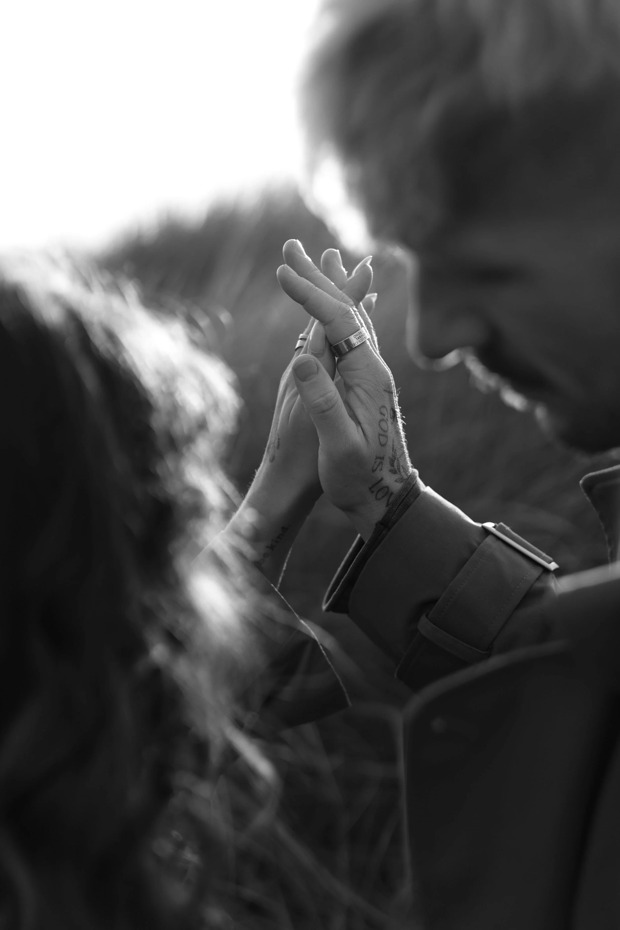 Black and white photo of two people with intertwined hands in a prayer or intimate gesture, with one person's profile partially visible and the other person's face blurred. The scene appears to be outdoors with soft lighting.