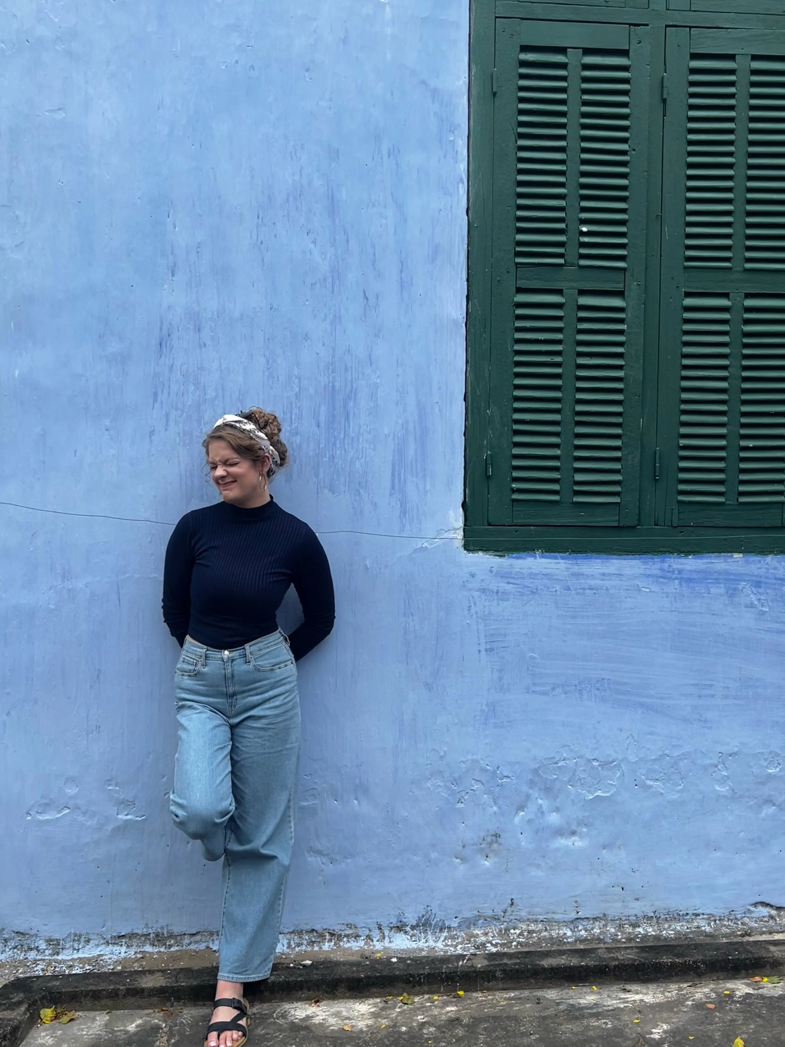 A woman standing against a blue wall with a green window shutter, smiling and wearing a black top, light blue jeans, and black sandals.