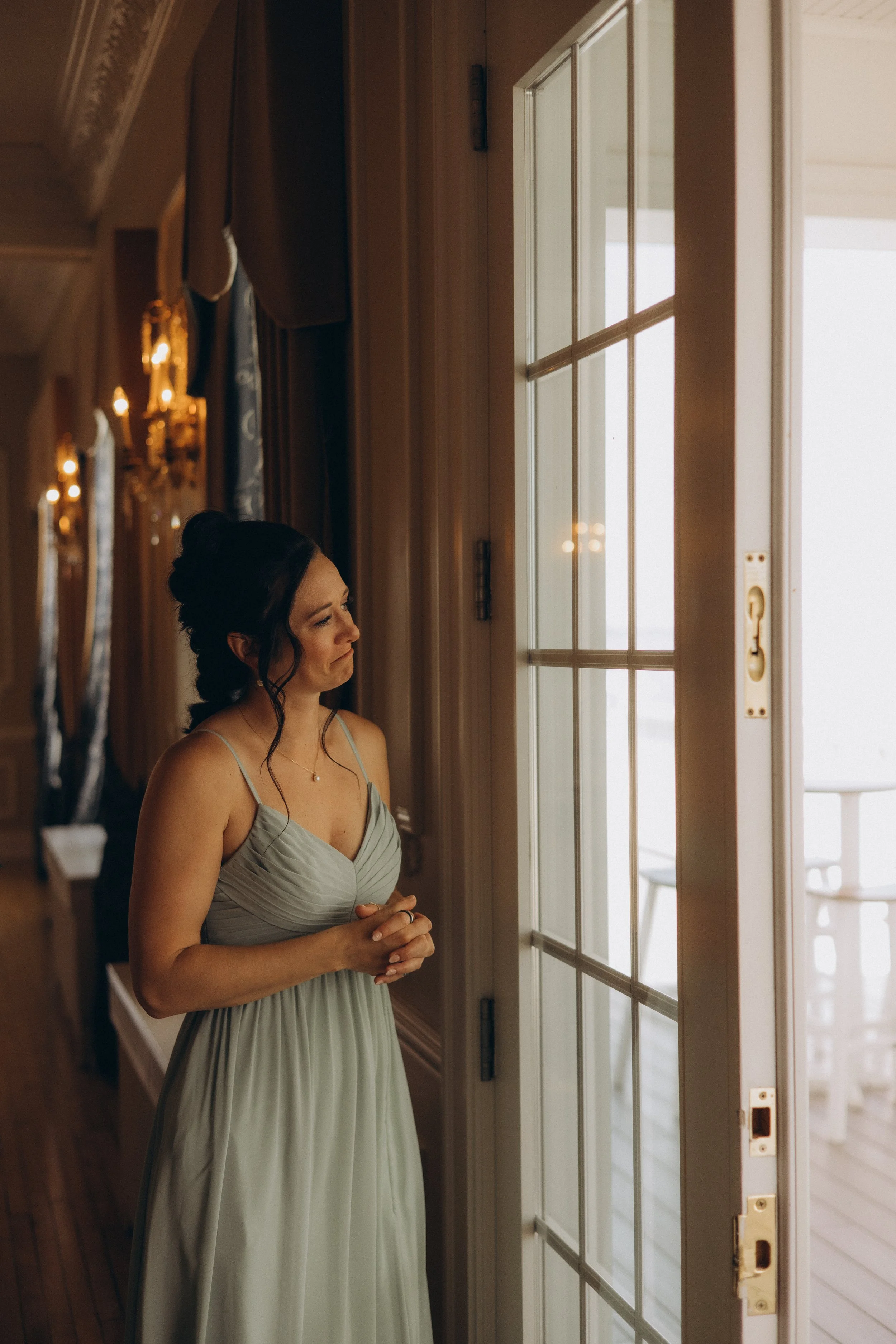 A woman with dark hair styled in an elegant updo wearing a light-colored dress, standing indoors near a glass-paneled door, looking outside with a soft expression.