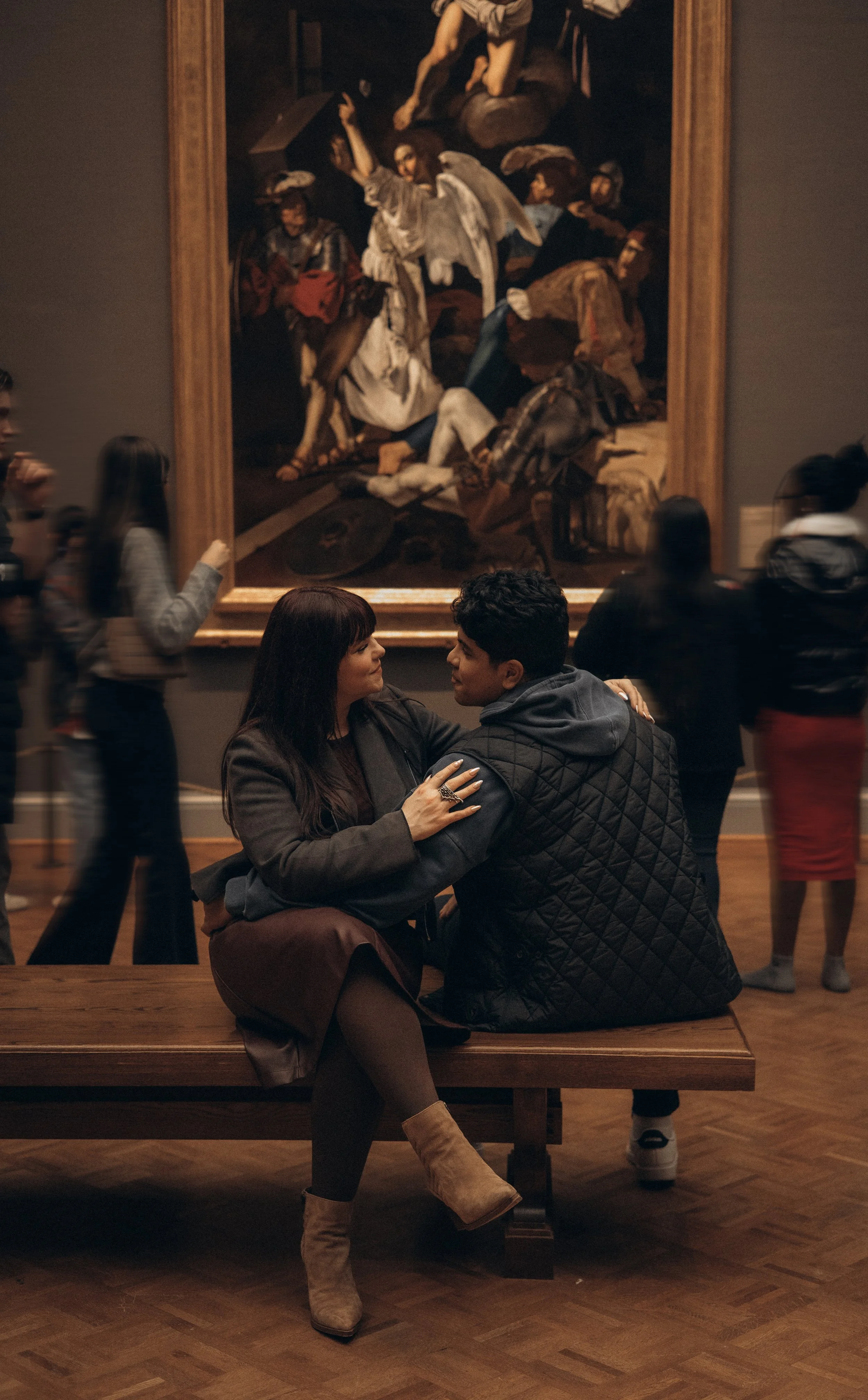 Two people, a woman and a man, sitting on a wooden bench in an art gallery, dancing and looking at each other. Behind them is a large painting depicting a biblical scene with several figures.