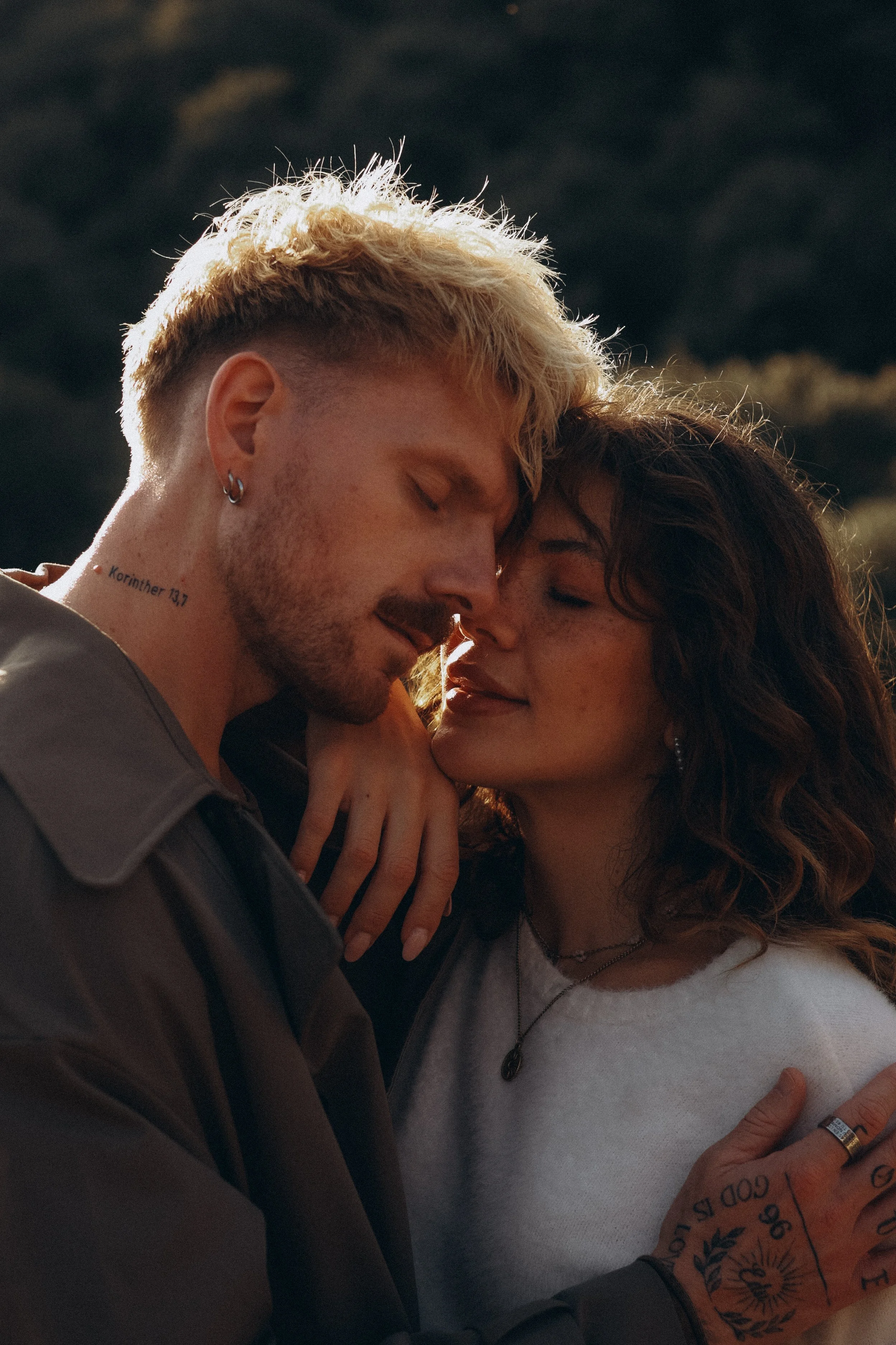 A close-up photo of a couple touching forehead and eyes closed, showing intimacy in an outdoor setting with trees in the background.