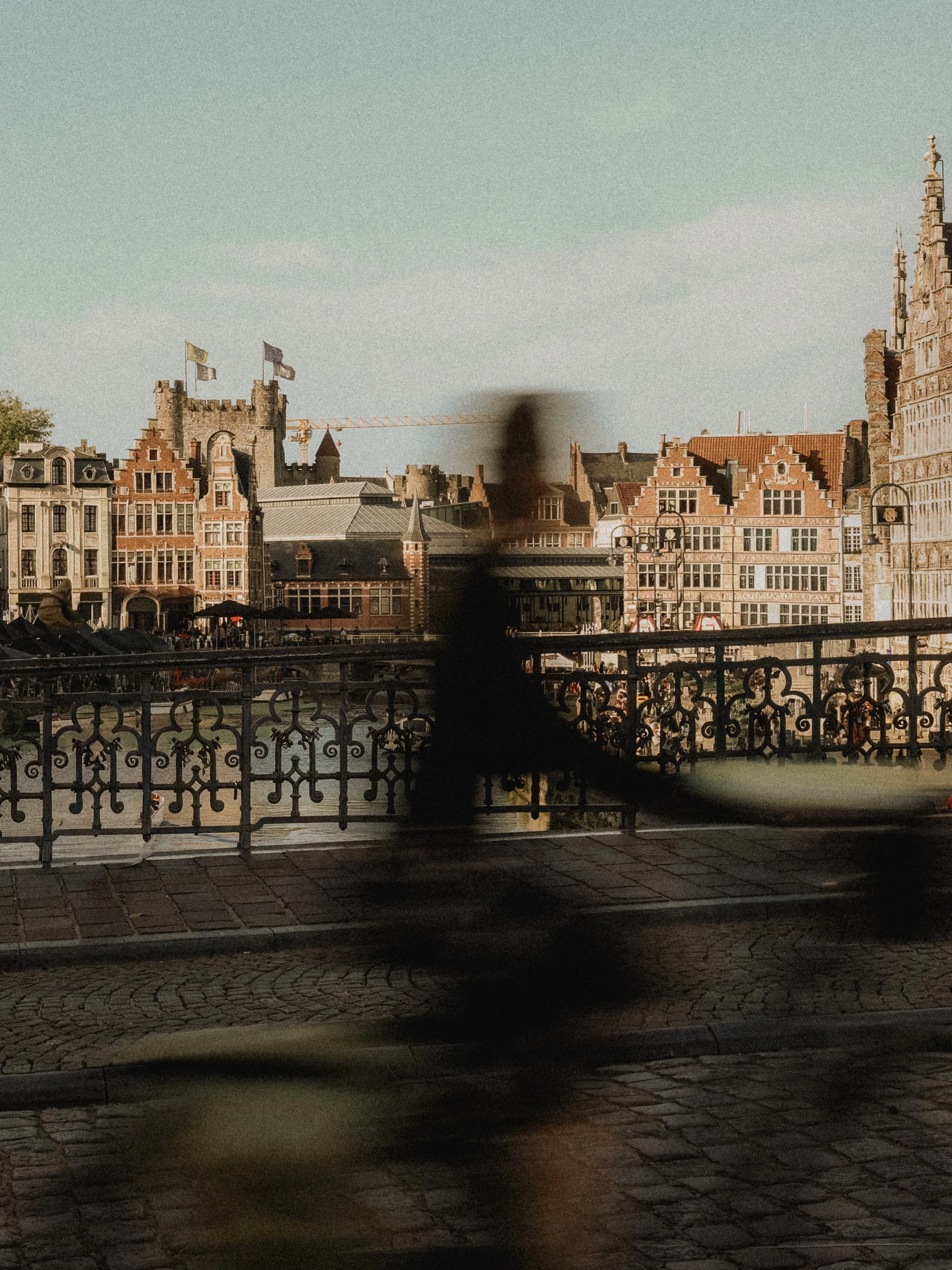 Cityscape view with historic European-style buildings, a construction crane, and a partly cloudy sky in the background. Foreground shows a decorative bridge railing and blurred figures.