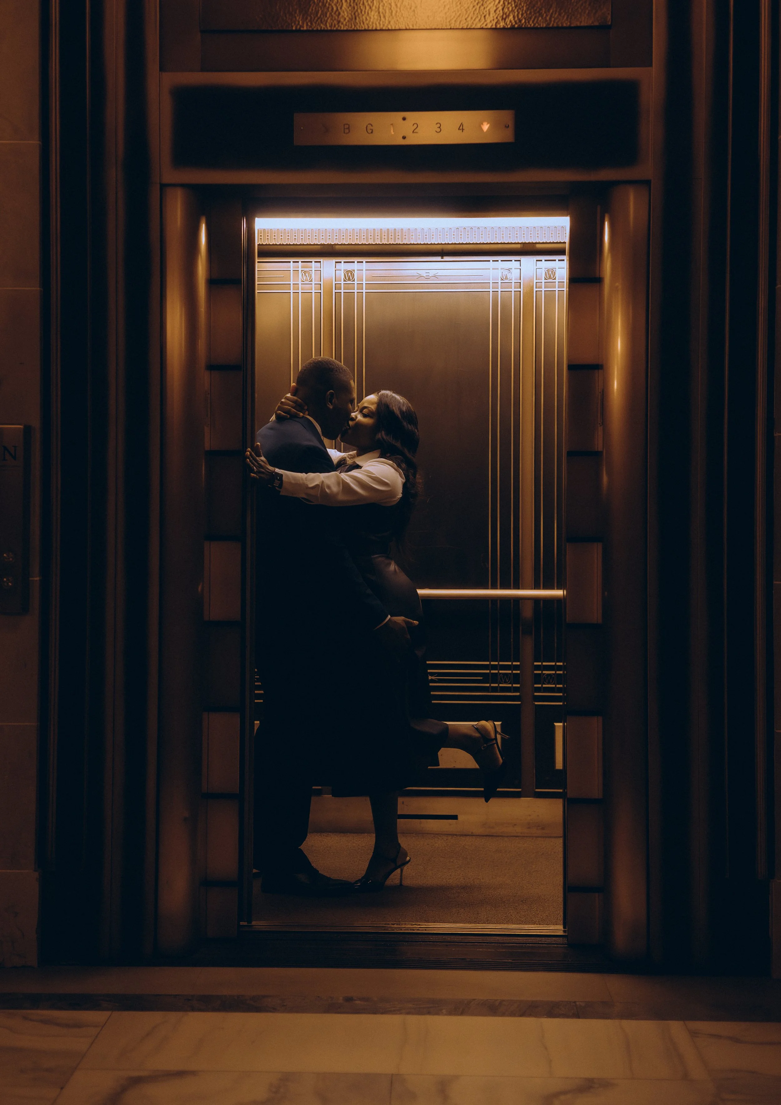 A couple sharing a kiss inside an elevator with warm lighting, with the woman on her knees embracing the man and wearing high heels.