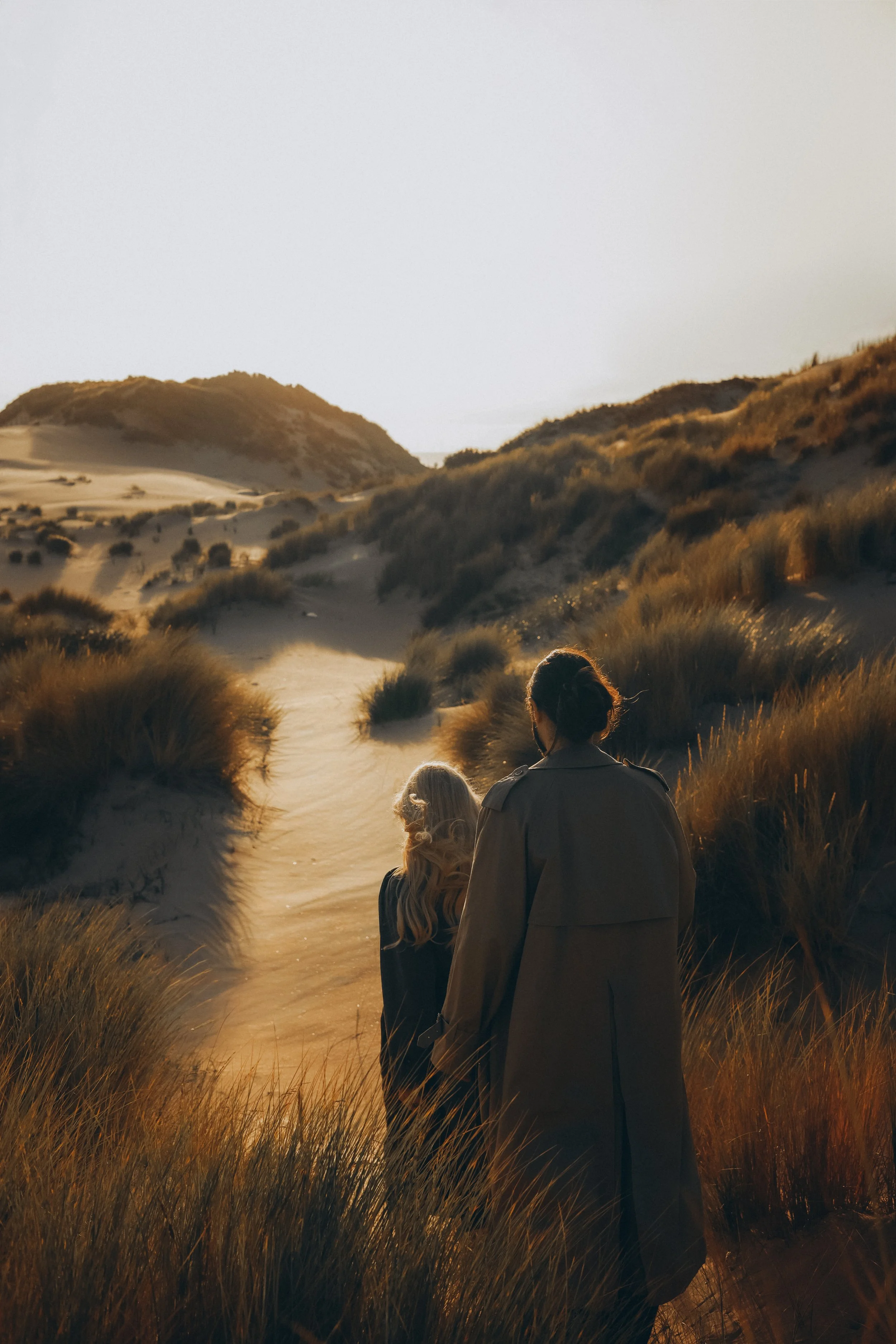 Two women walking along a sandy trail through a desert landscape with hills and dry vegetation during sunset.