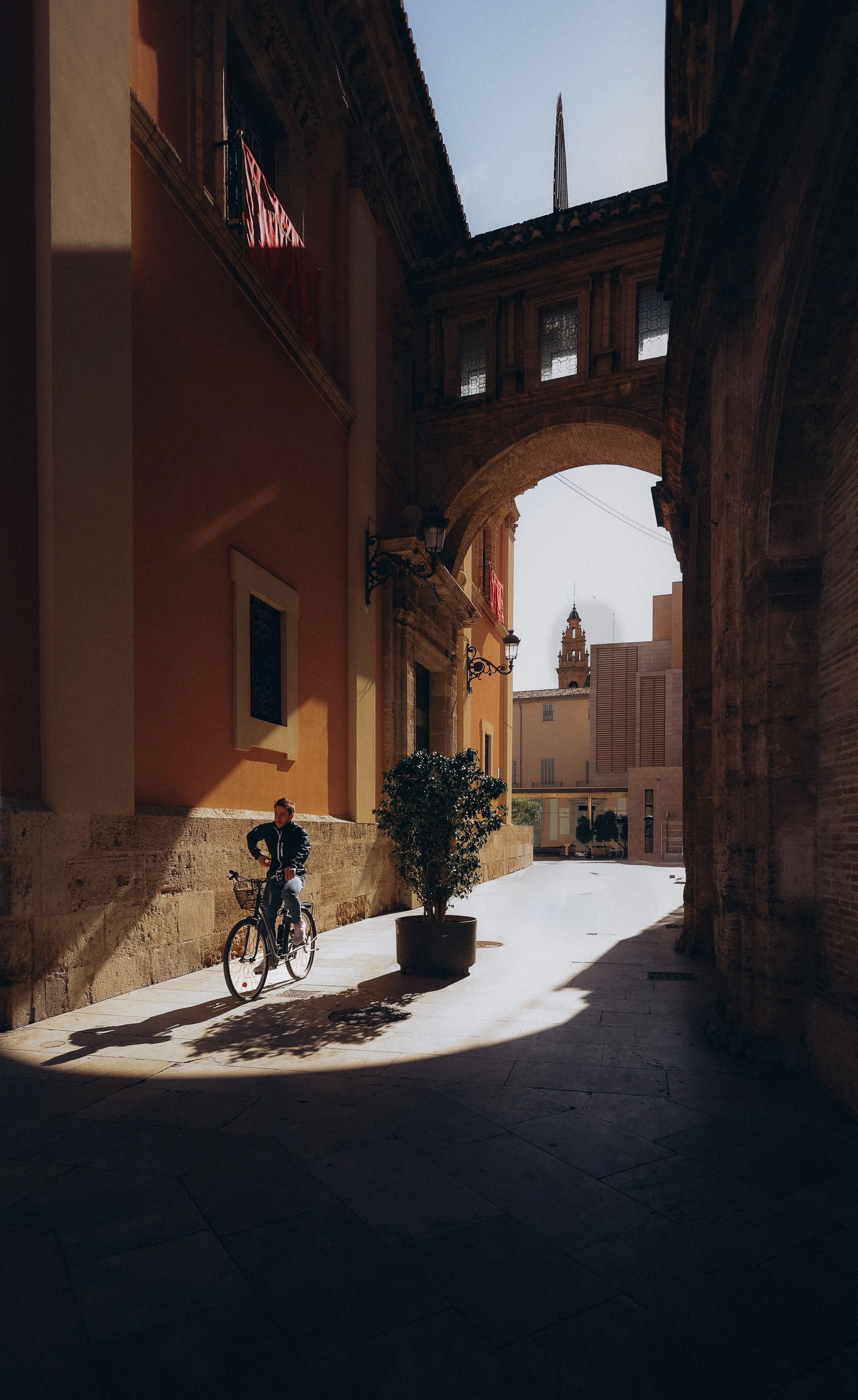 A person riding a bicycle in a shadowed street area with buildings and arches, and a church tower visible in the background.