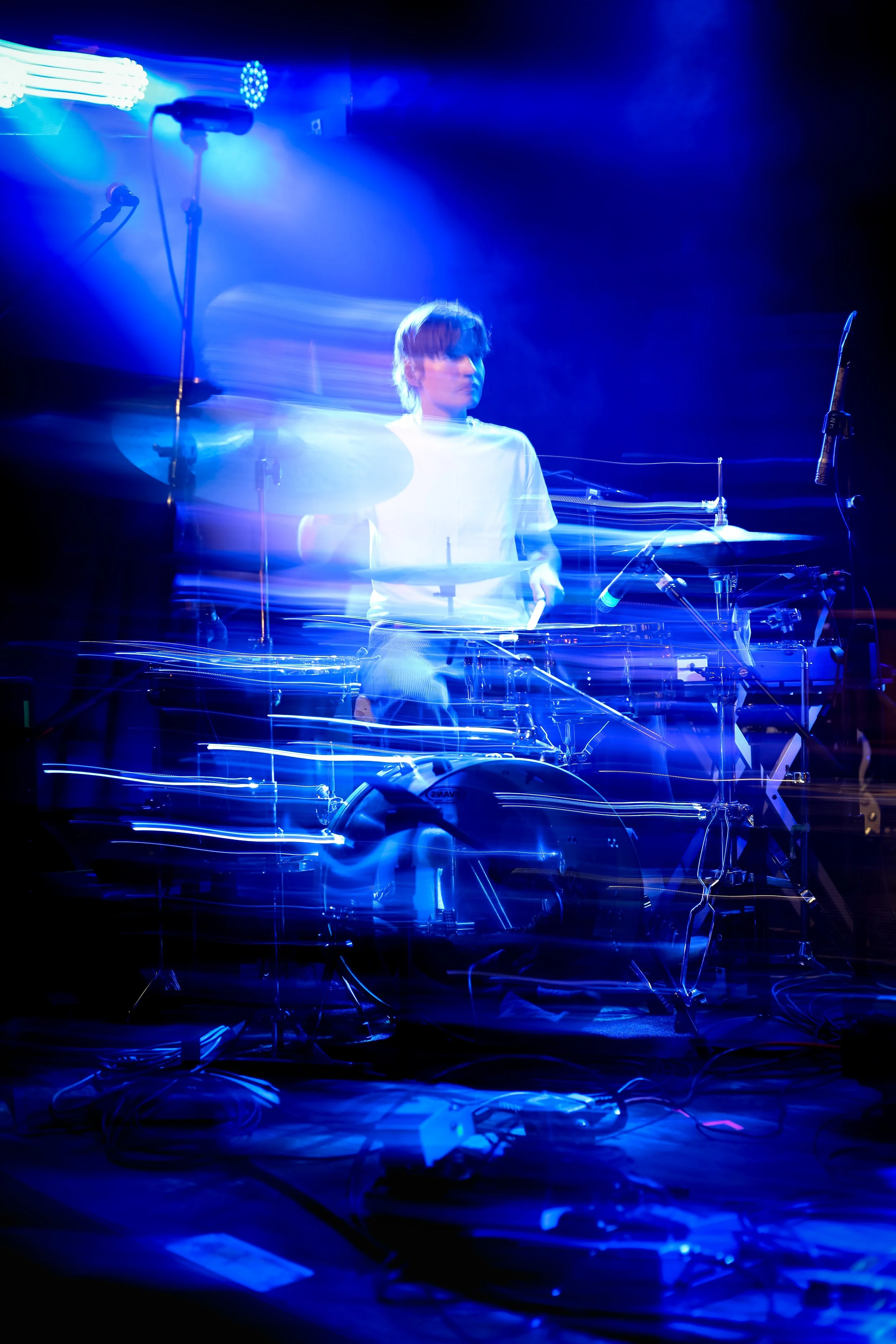 A person appears to be at a stage with blue lighting, sitting behind electronic music equipment with various wires and microphones, creating a motion blur effect.