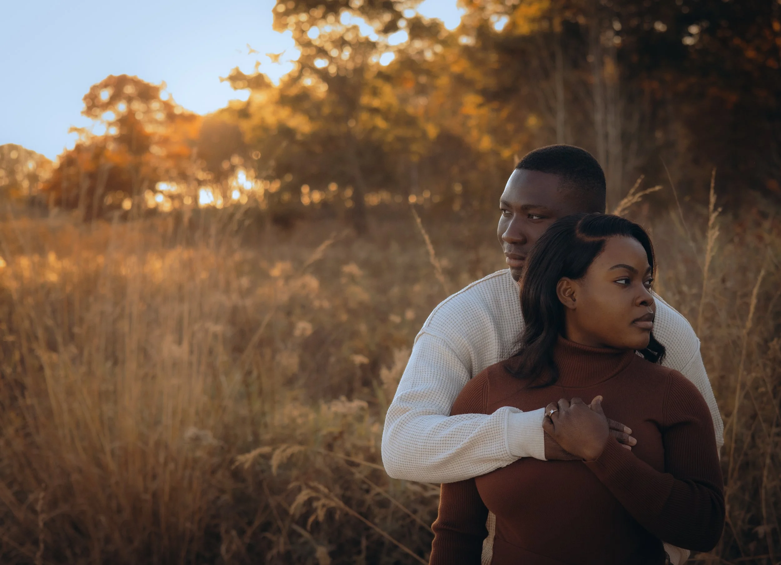 A man and woman embrace outdoors during sunset, with trees and tall grass in the background.